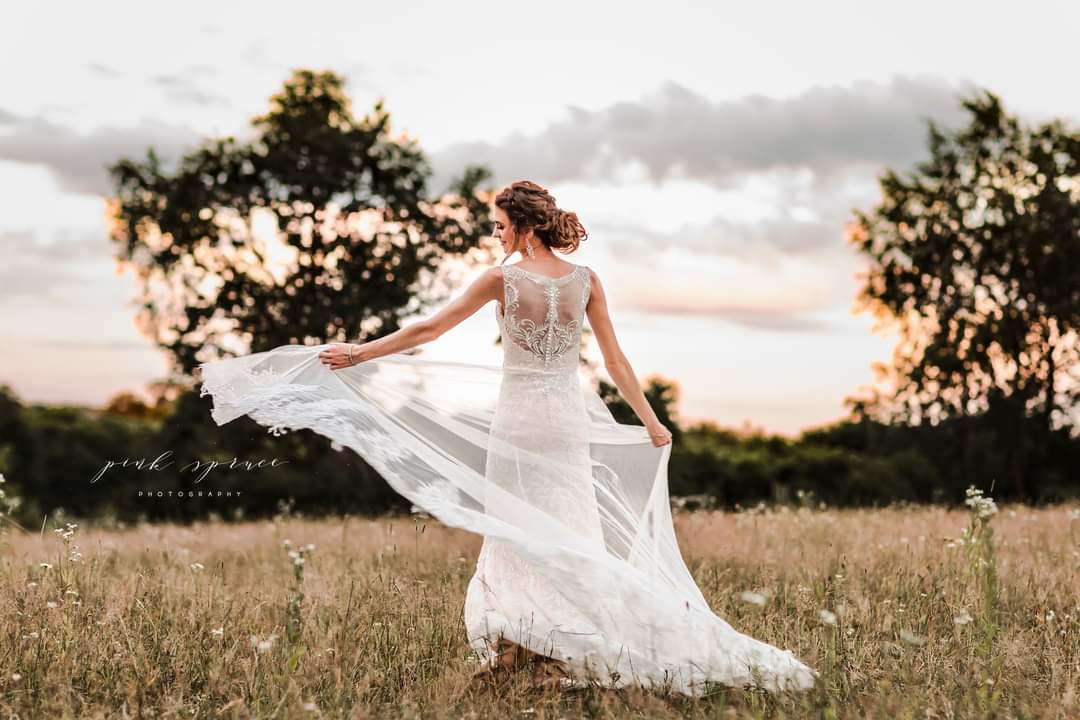 Woman in a wedding dress standing in a field during sunset, holding her dress out to the side.