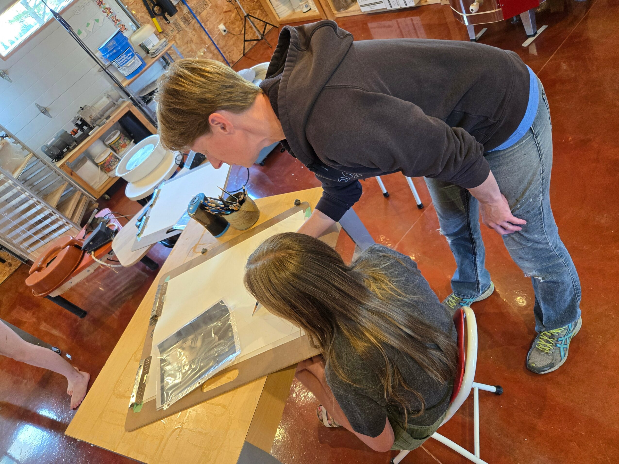 Two people, a woman and a girl, are leaning over a table looking at a book or magazine. The woman has short blonde hair and is wearing a black hoodie. The girl has long brown hair and is wearing a dark top. The setting appears to be a room with wooden floors and various objects in the background.