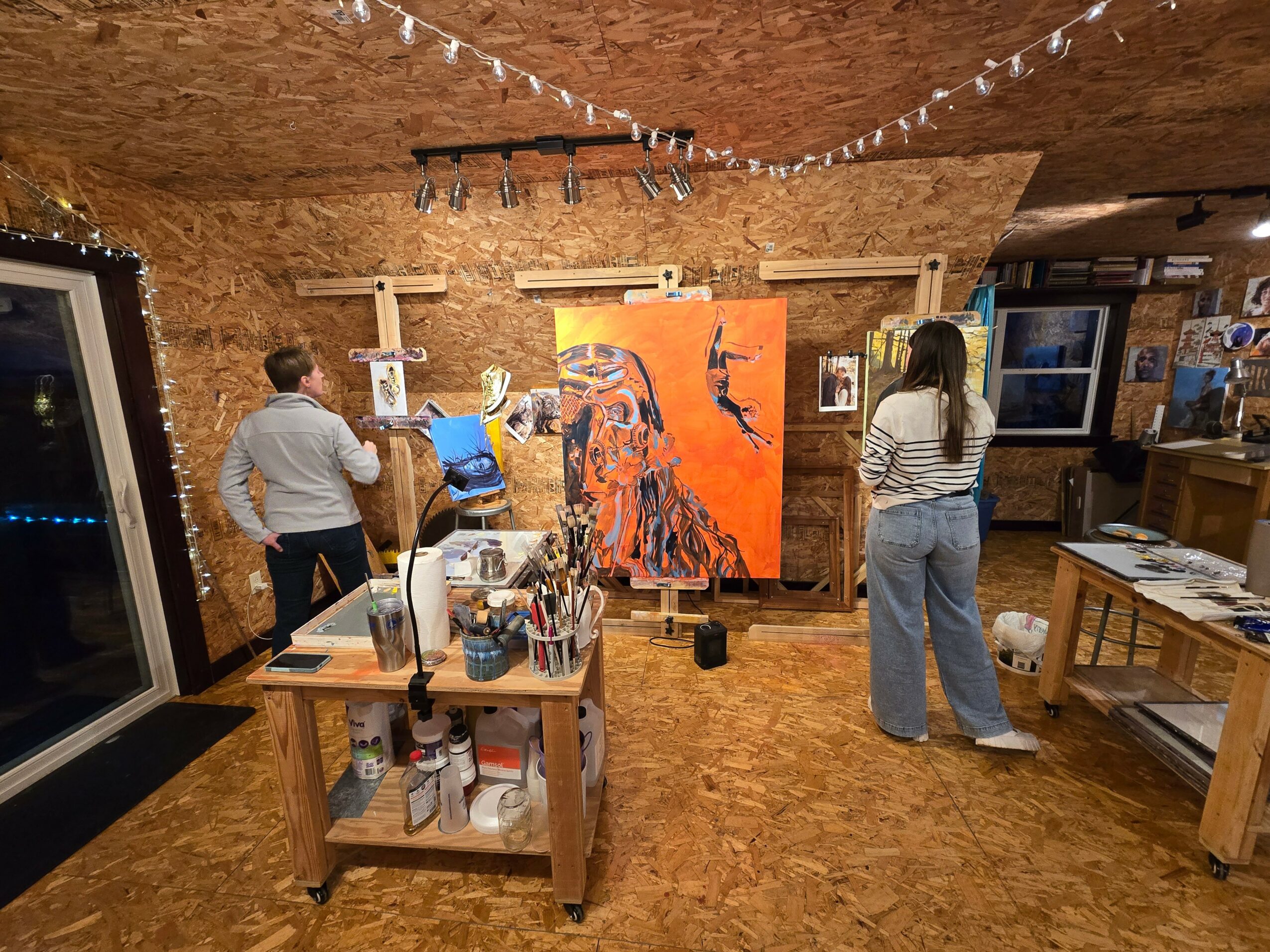 Two people observe a large painting of a woman with a bird on her head in an art studio with wooden walls and ceiling.