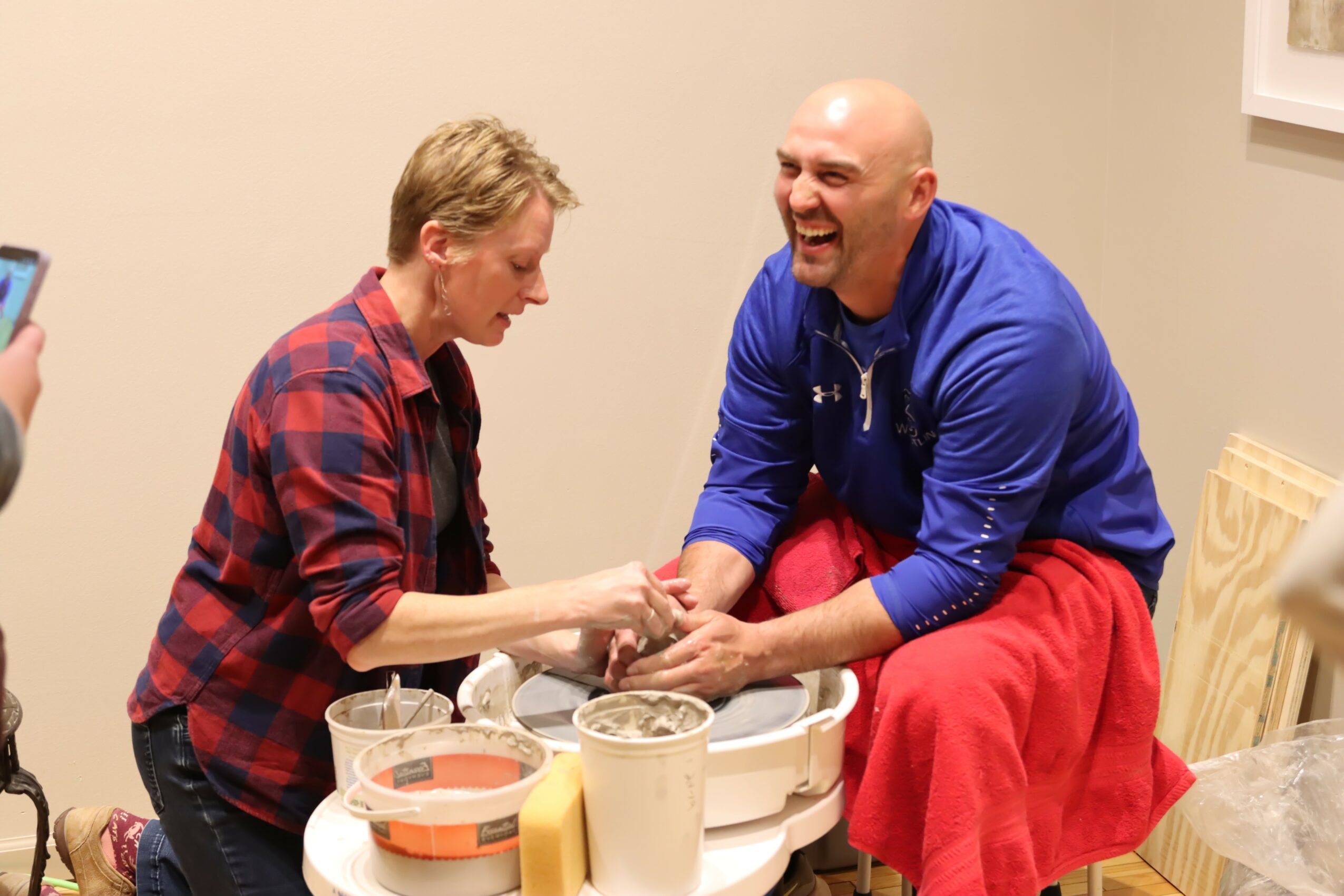 Two men sit and laugh, one in a red plaid shirt and the other in a blue jacket, holding hands.