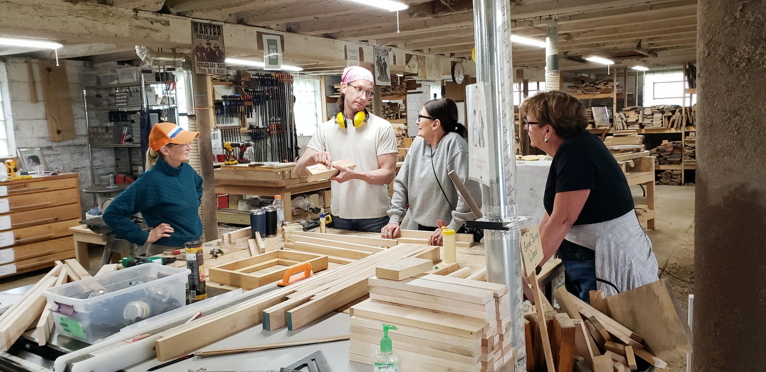 Four people working with wood in a workshop, surrounded by woodworking tools and materials.