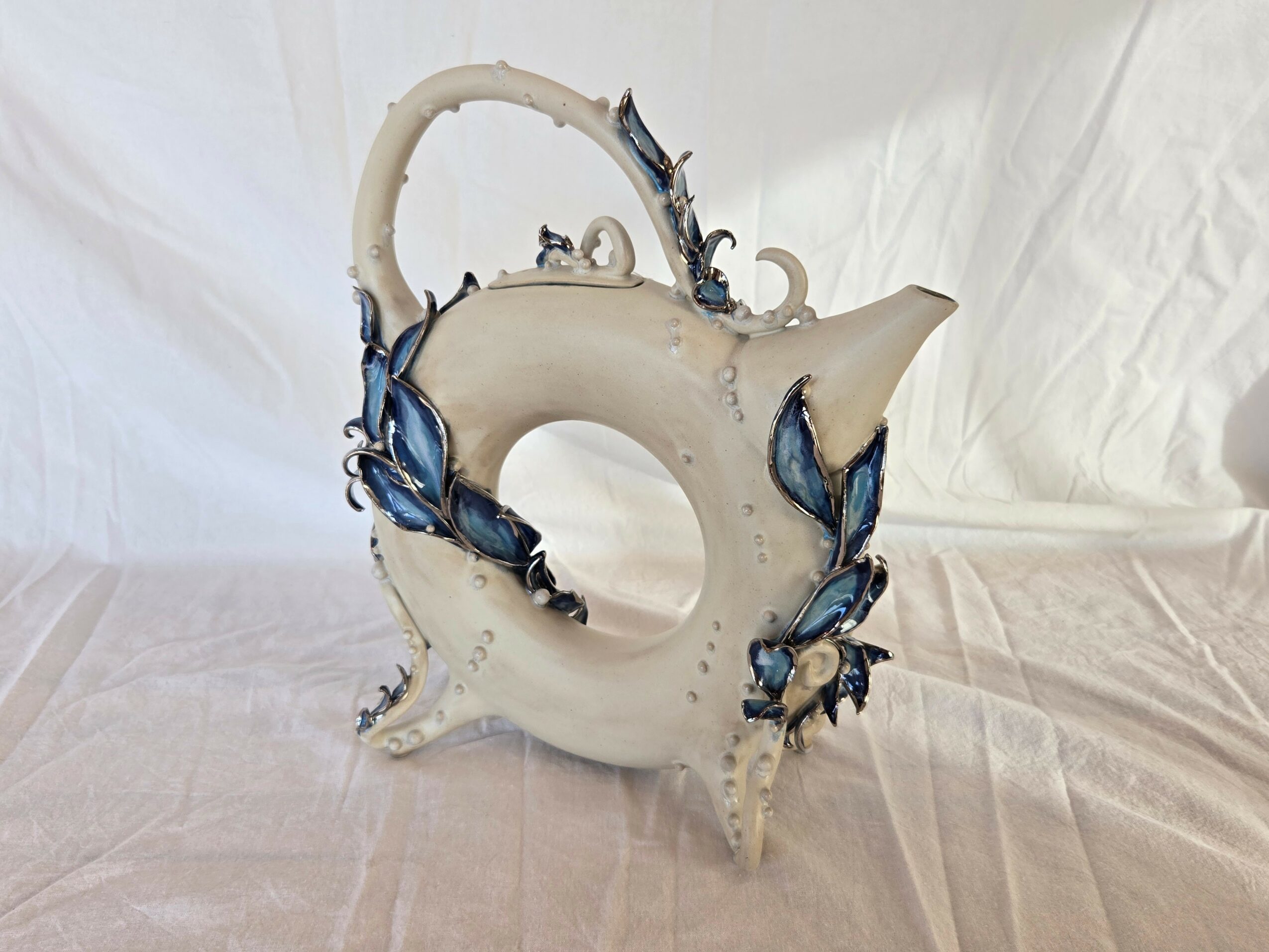 Decorative ceramic teapot with blue floral accents, curved handle, and ornate design, placed on a fabric surface with a plain background.