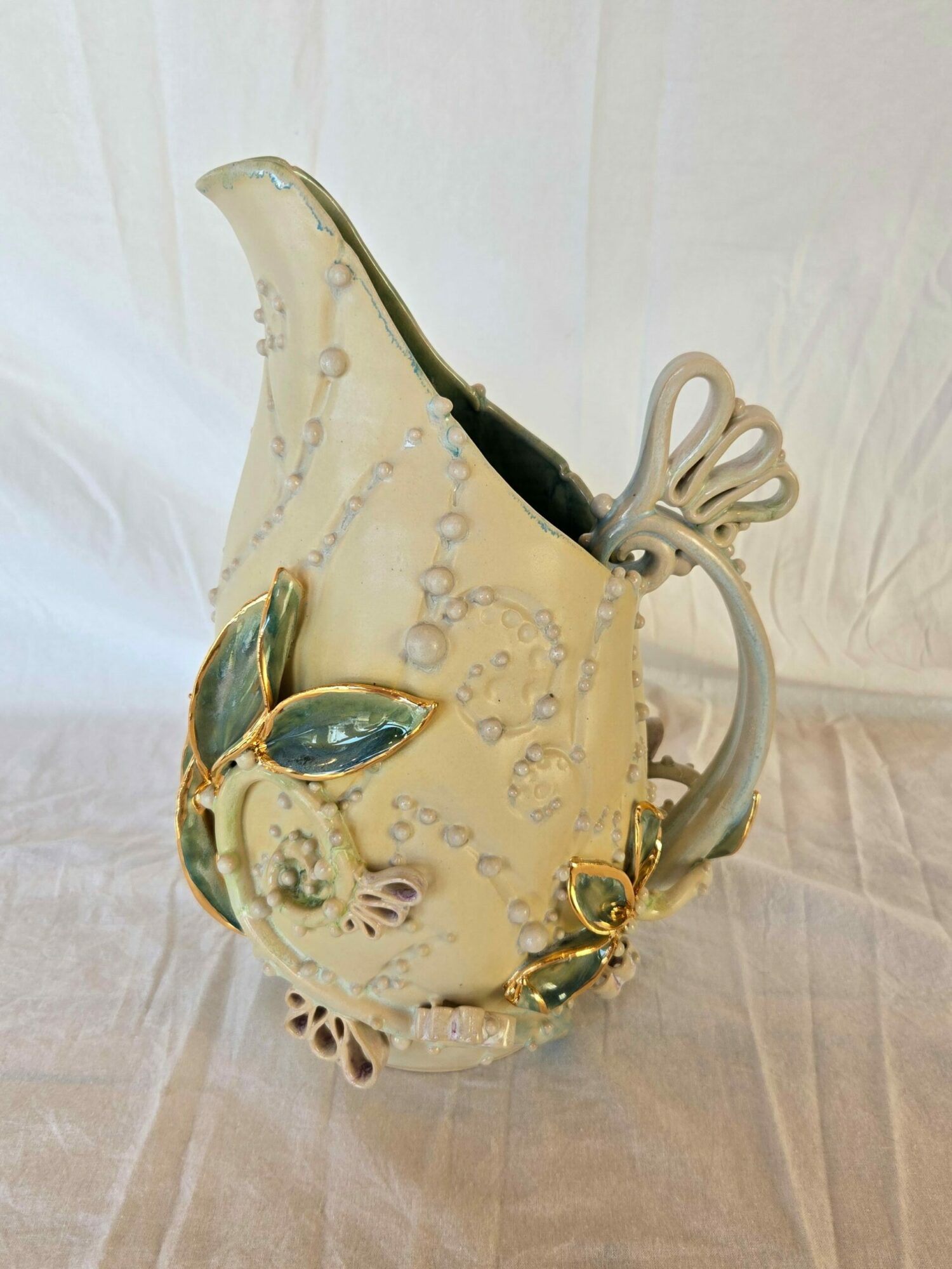 Decorative glass teapot with gold accents and blue details, viewed from the side, on a fabric background.
