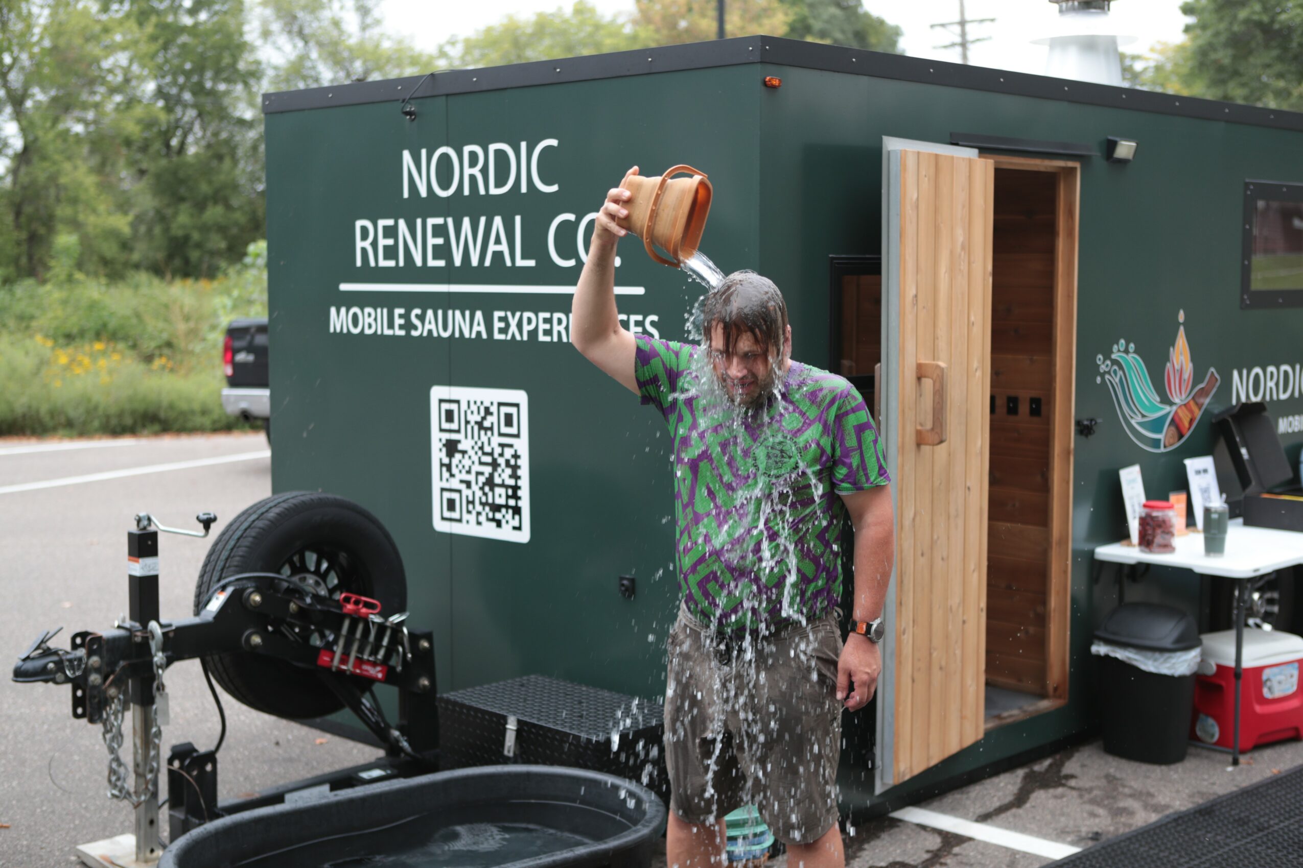 Person with wet hair and clothes standing outside a green mobile sauna, pouring water over their head, with trees in background.