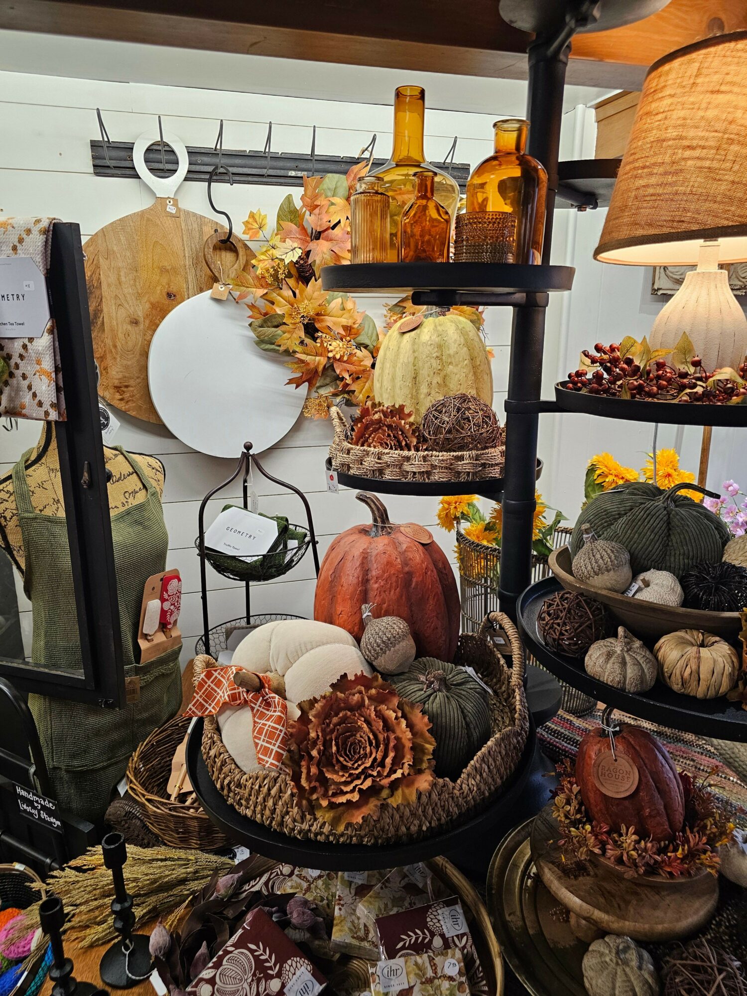 Decorative fall-themed display with pumpkins, flowers, and candles on a black metal stand, with a white plate and a lamp nearby.