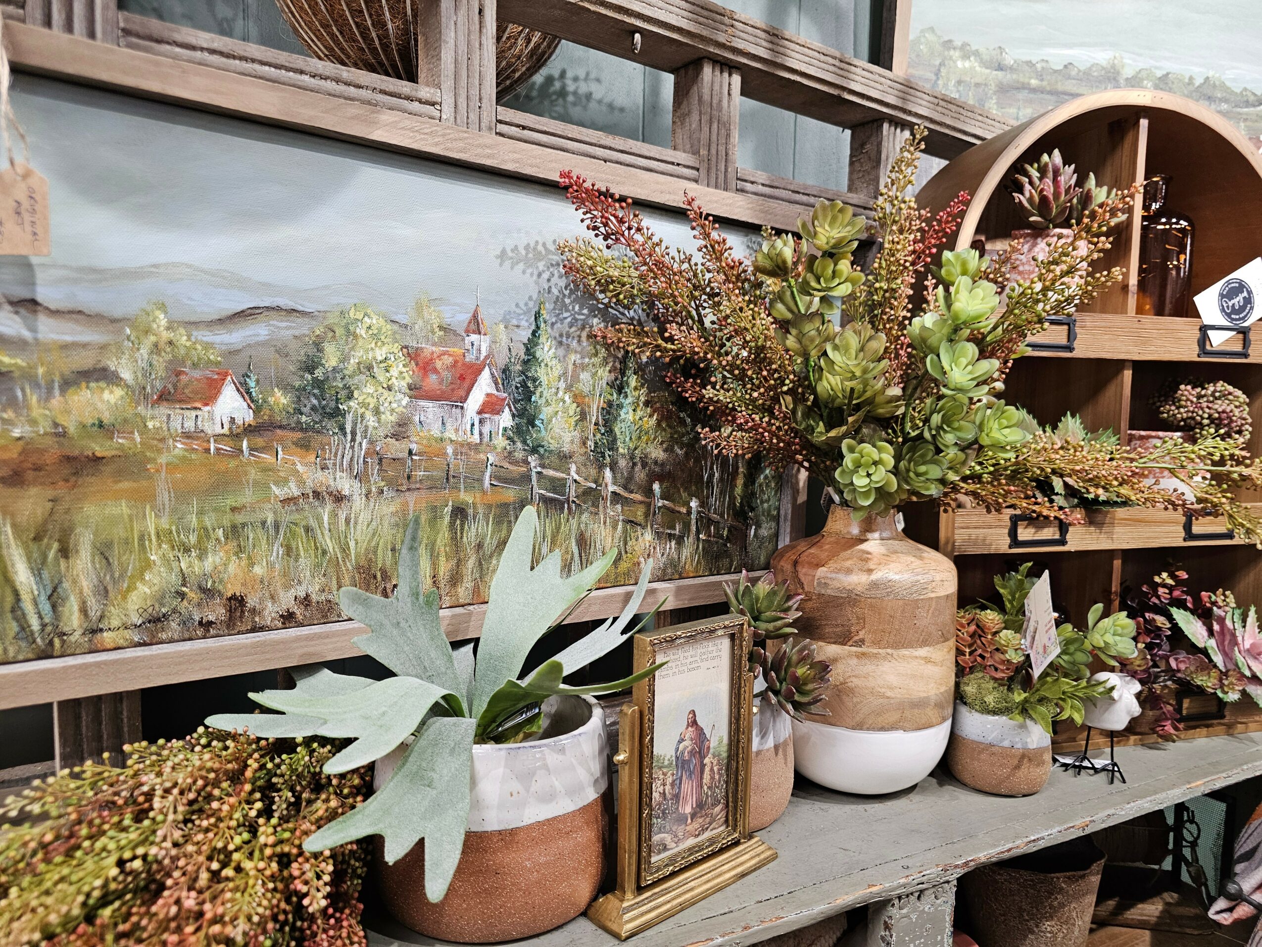 Display of potted plants, a framed picture, and decorative items on a shelf with a landscape painting in the background.