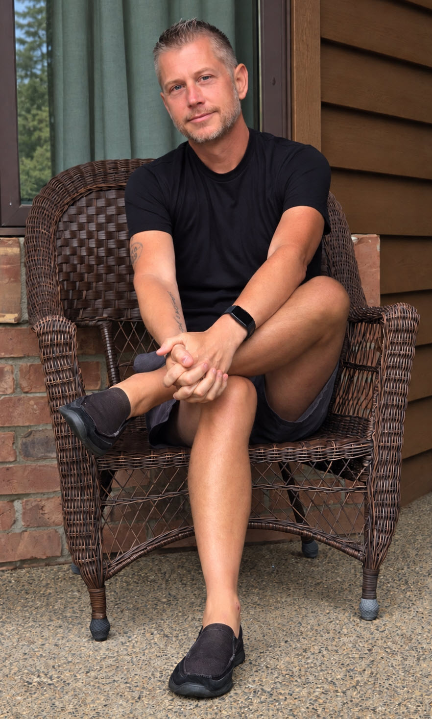 Person sitting on a wicker chair outdoors, wearing a black t-shirt and shorts, with arms crossed and legs crossed.