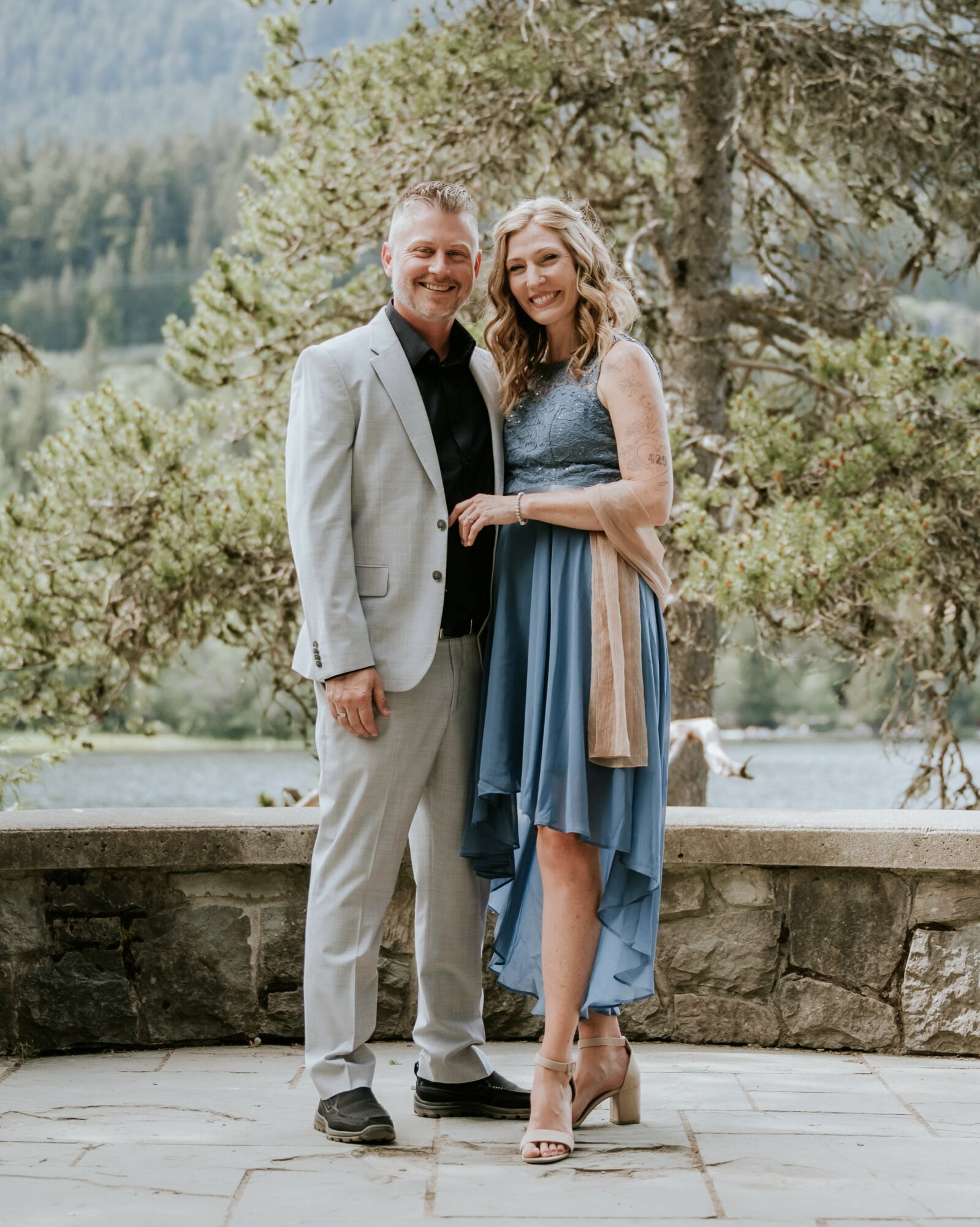 A smiling couple stands outdoors near a stone wall with trees and a lake in the background.