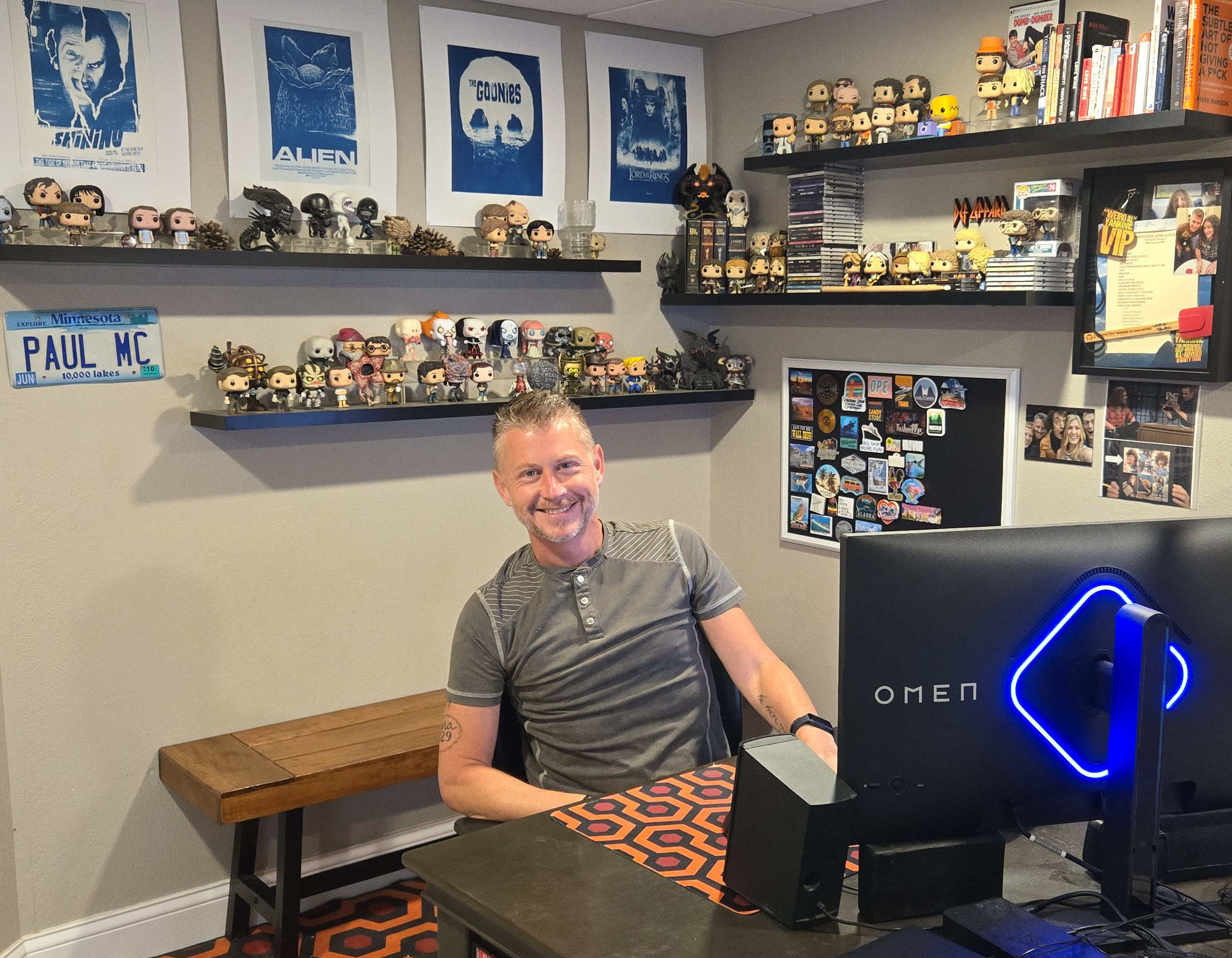 Man sitting at desk in room with shelves of collectibles and posters on wall, smiling at camera.
