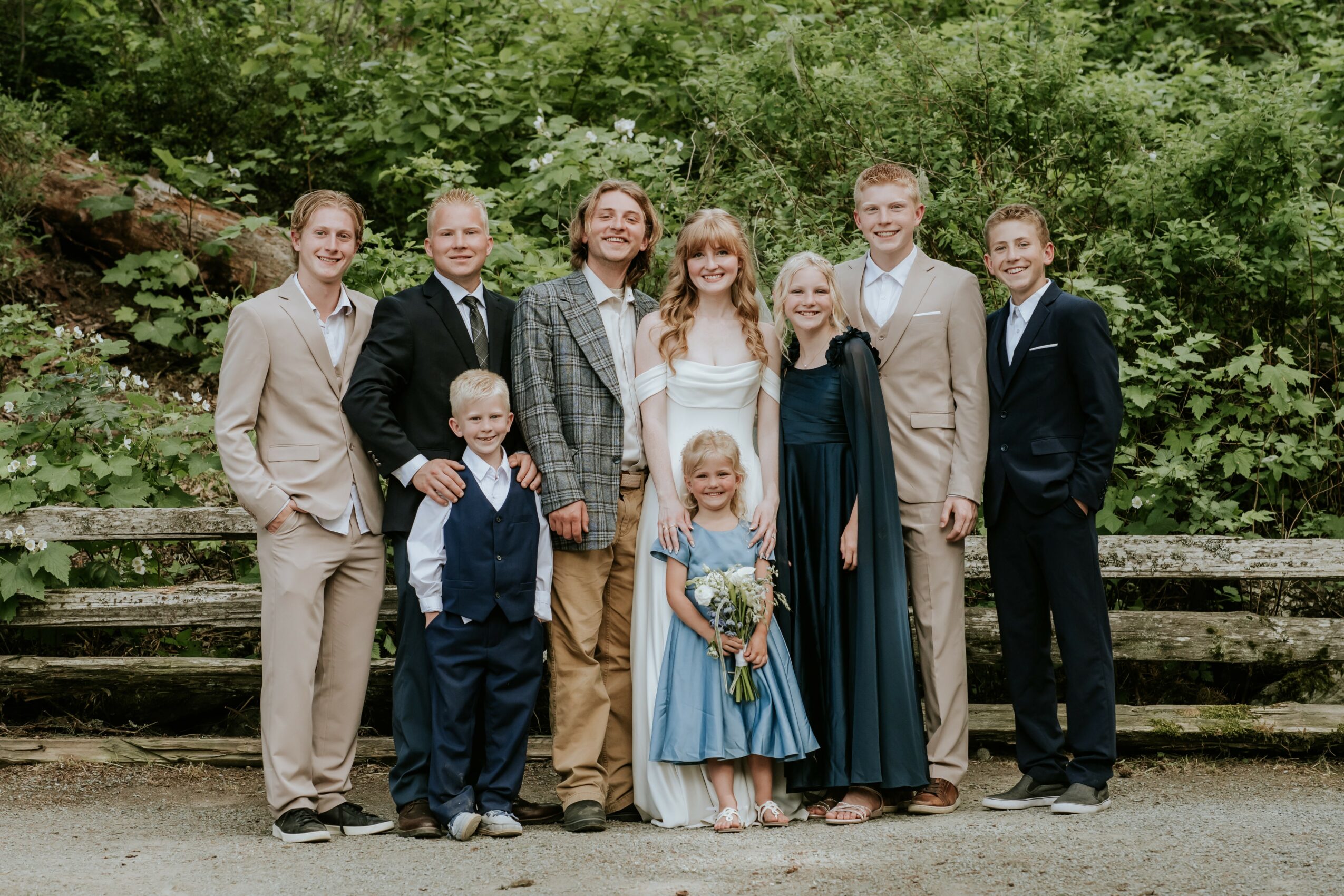Group of nine people, including children, standing outdoors on wooden steps with green trees in background.