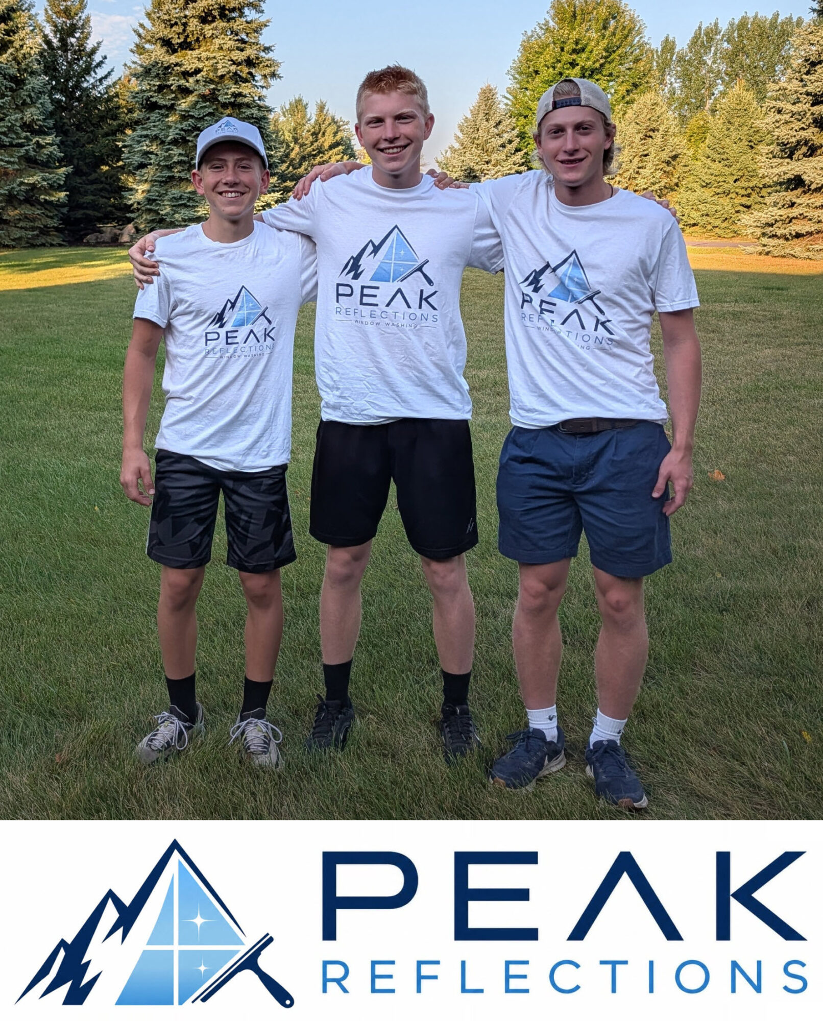 Three young men stand outdoors on grass, smiling, wearing matching white T-shirts with a mountain logo and text, with trees in background.