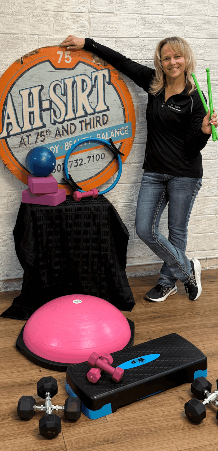 Woman standing next to a large circular sign with a pink bowling ball and equipment on the floor, holding green sticks, smiling.