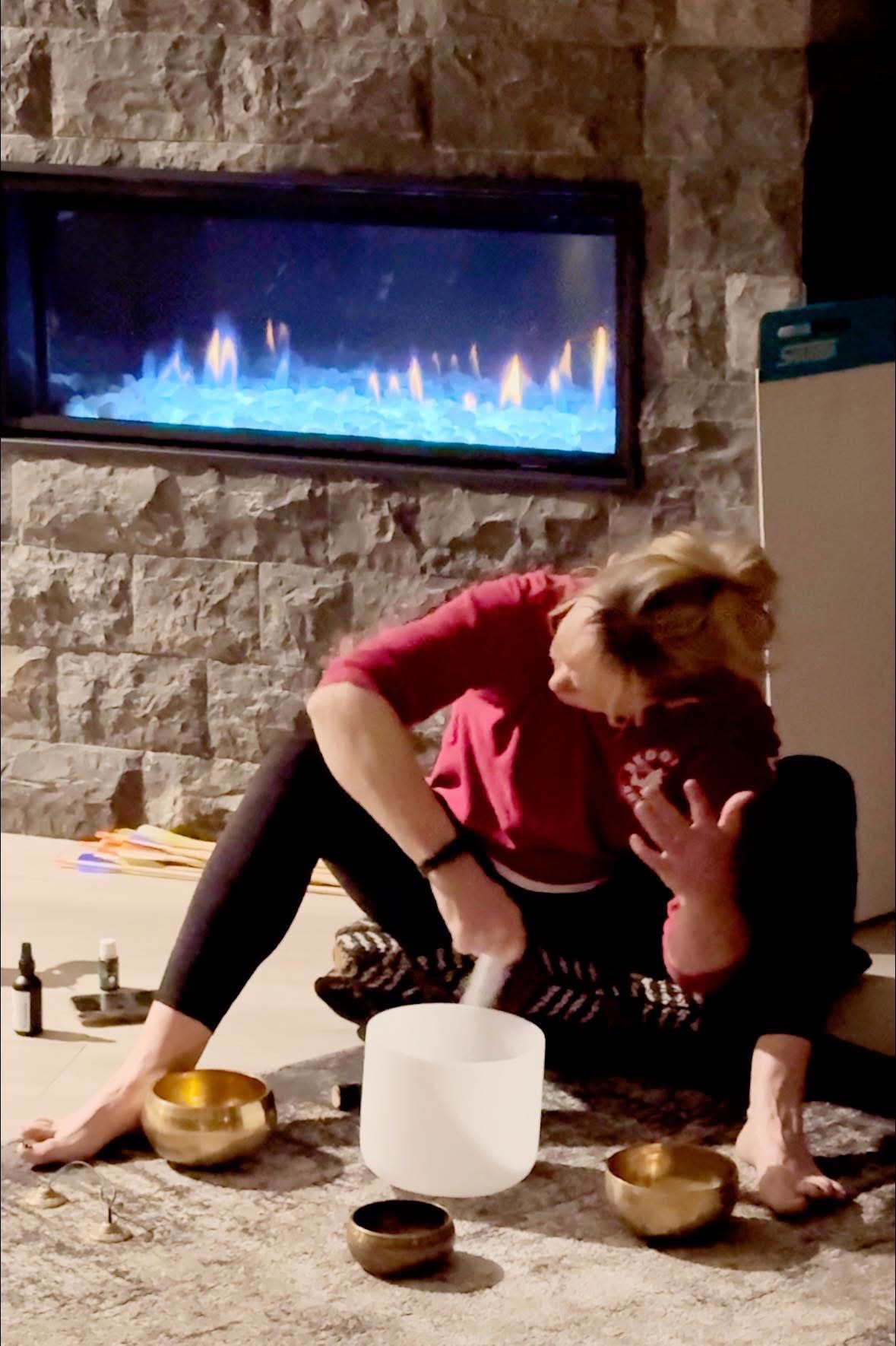 Person sitting on the floor with legs crossed, touching their face, in front of a fireplace with blue flames, on a stone wall background.