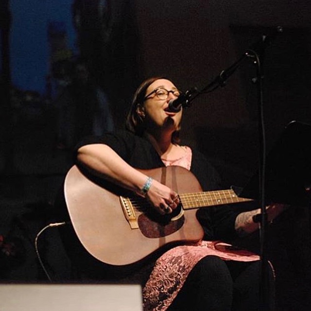 Person singing into microphone while playing an acoustic guitar, seated on stage with dark background.