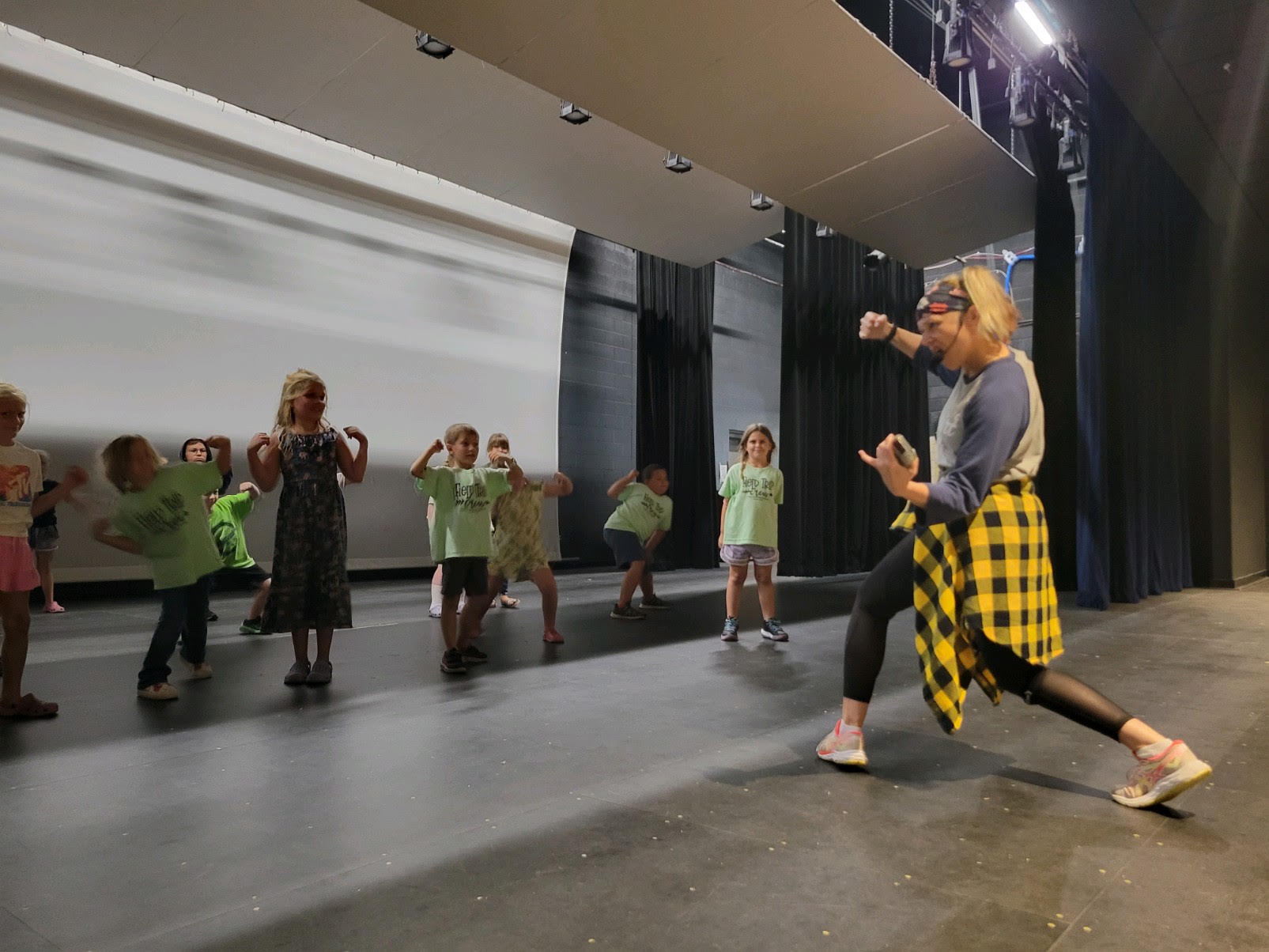 Children and an adult in a dance studio, practicing dance moves, with black curtains and a white wall in the background.