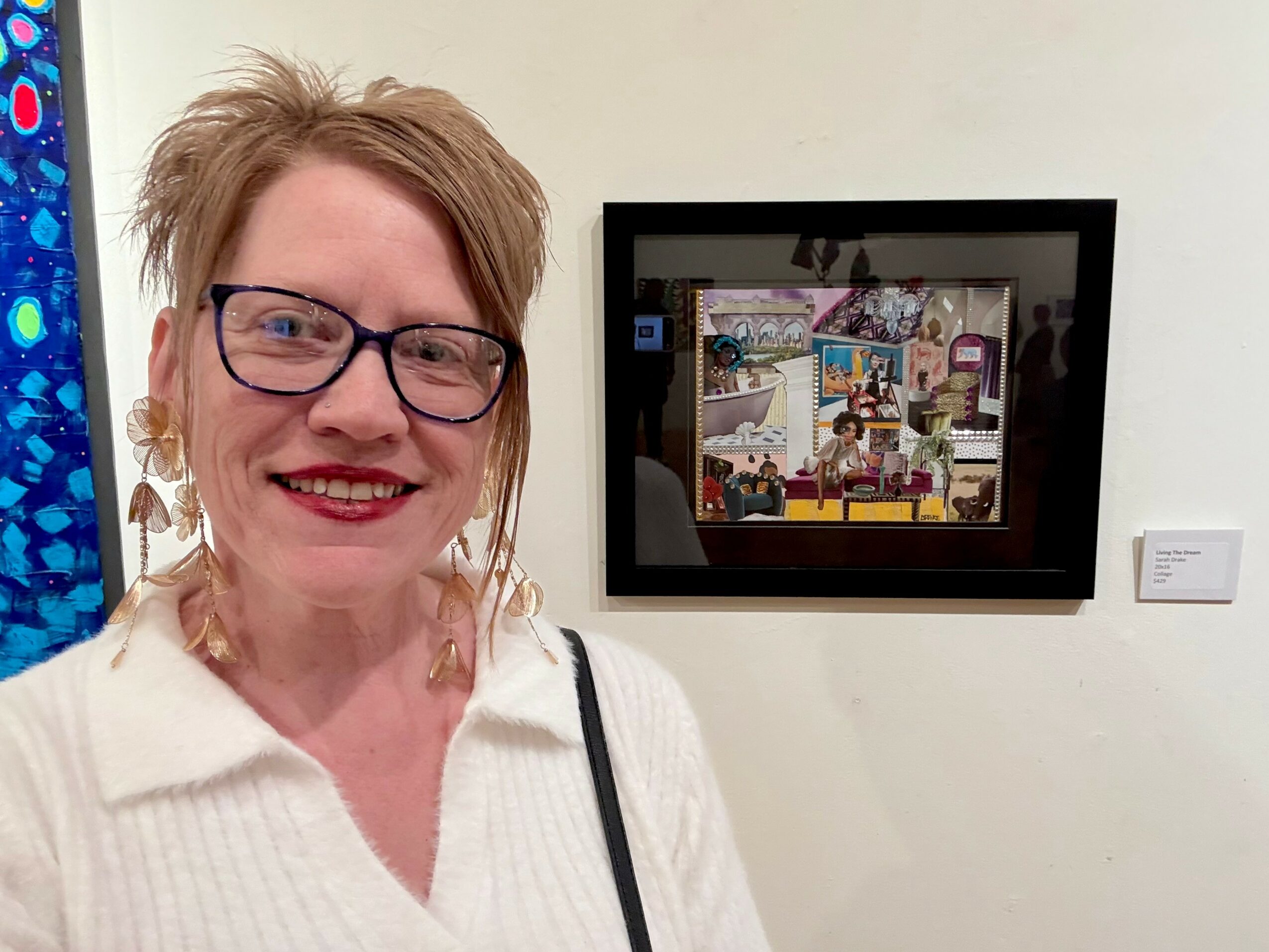 Smiling woman with glasses and earrings stands in front of a framed collage on a white wall.