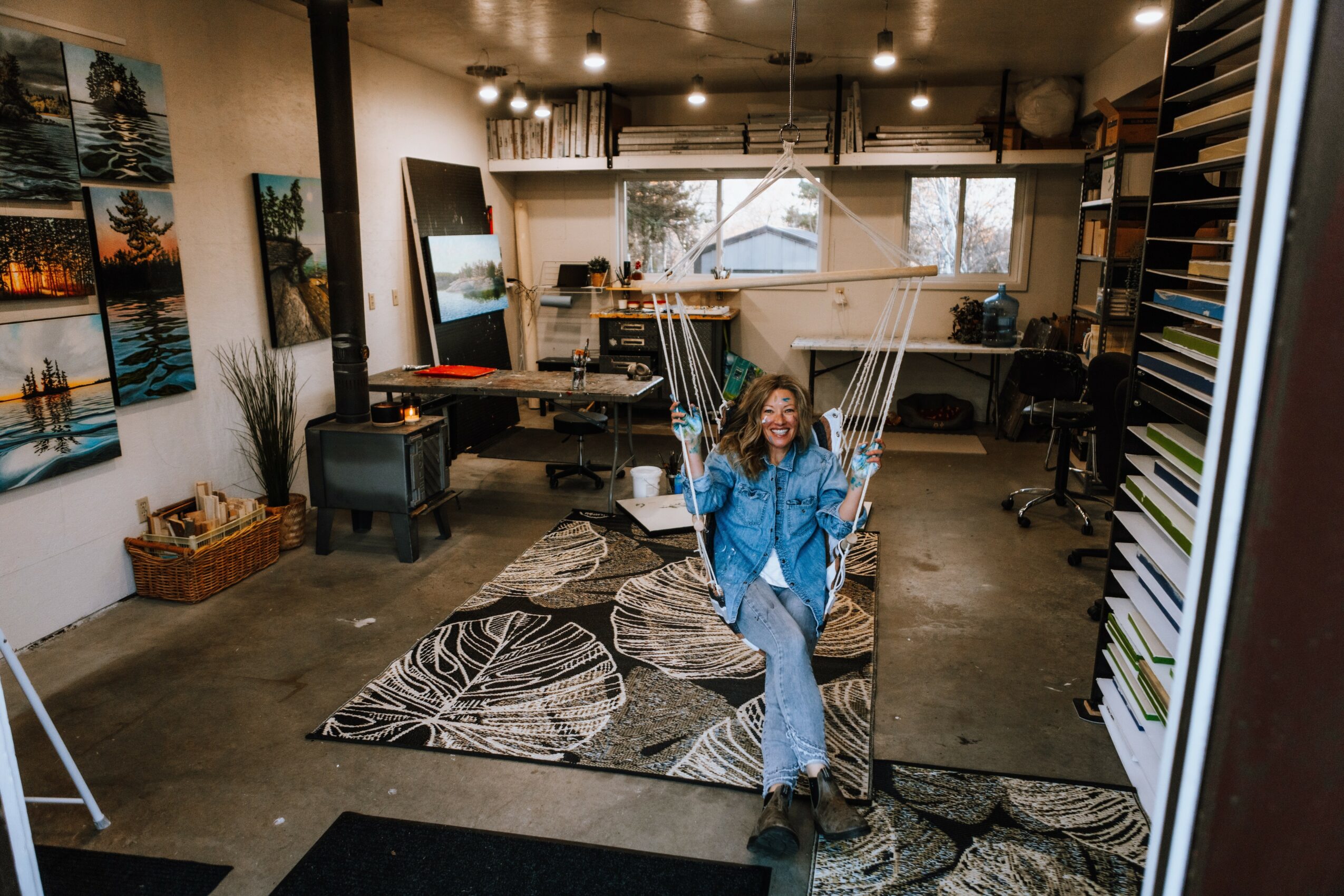 Woman in blue jacket sitting on a patterned rug in an art studio with paintings and supplies around.
