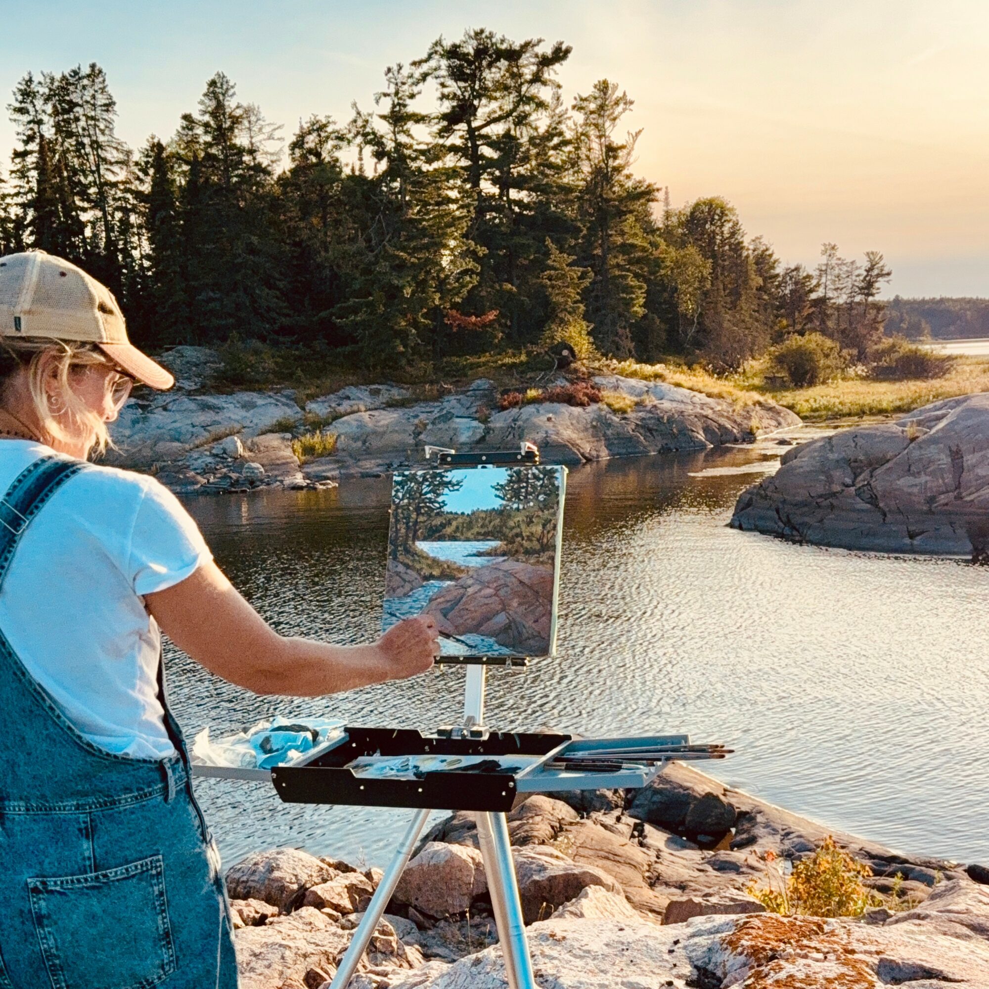 Person painting landscape by river at sunset, trees and rocks in background, wearing a cap and overalls.