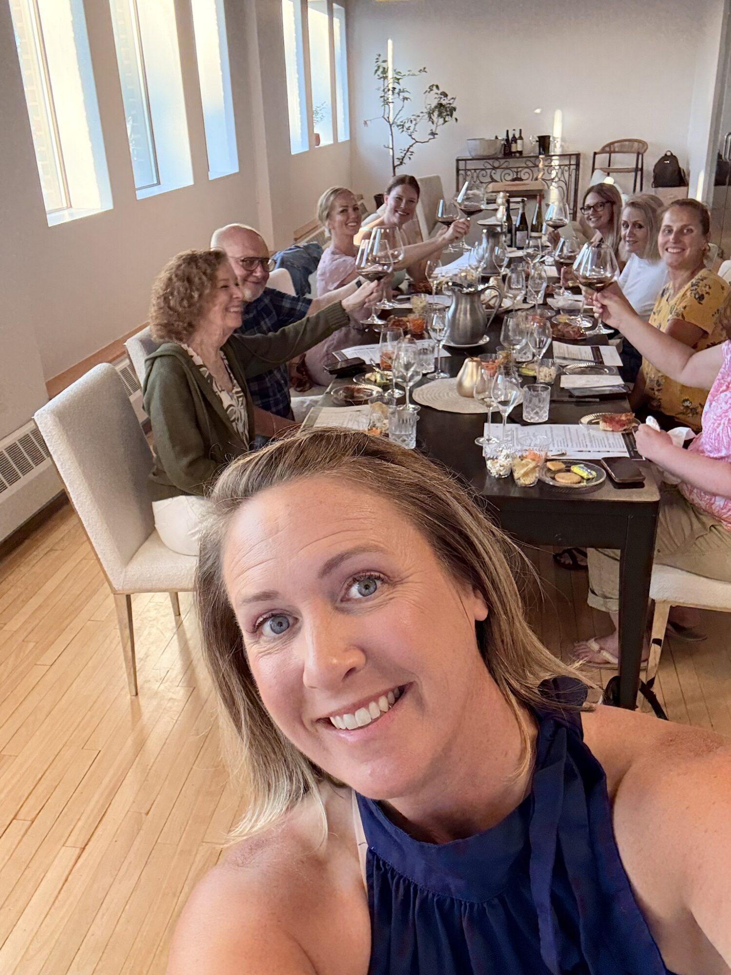 Group of people sitting at a long dining table, smiling and raising glasses, in a bright room with windows and wooden floors.