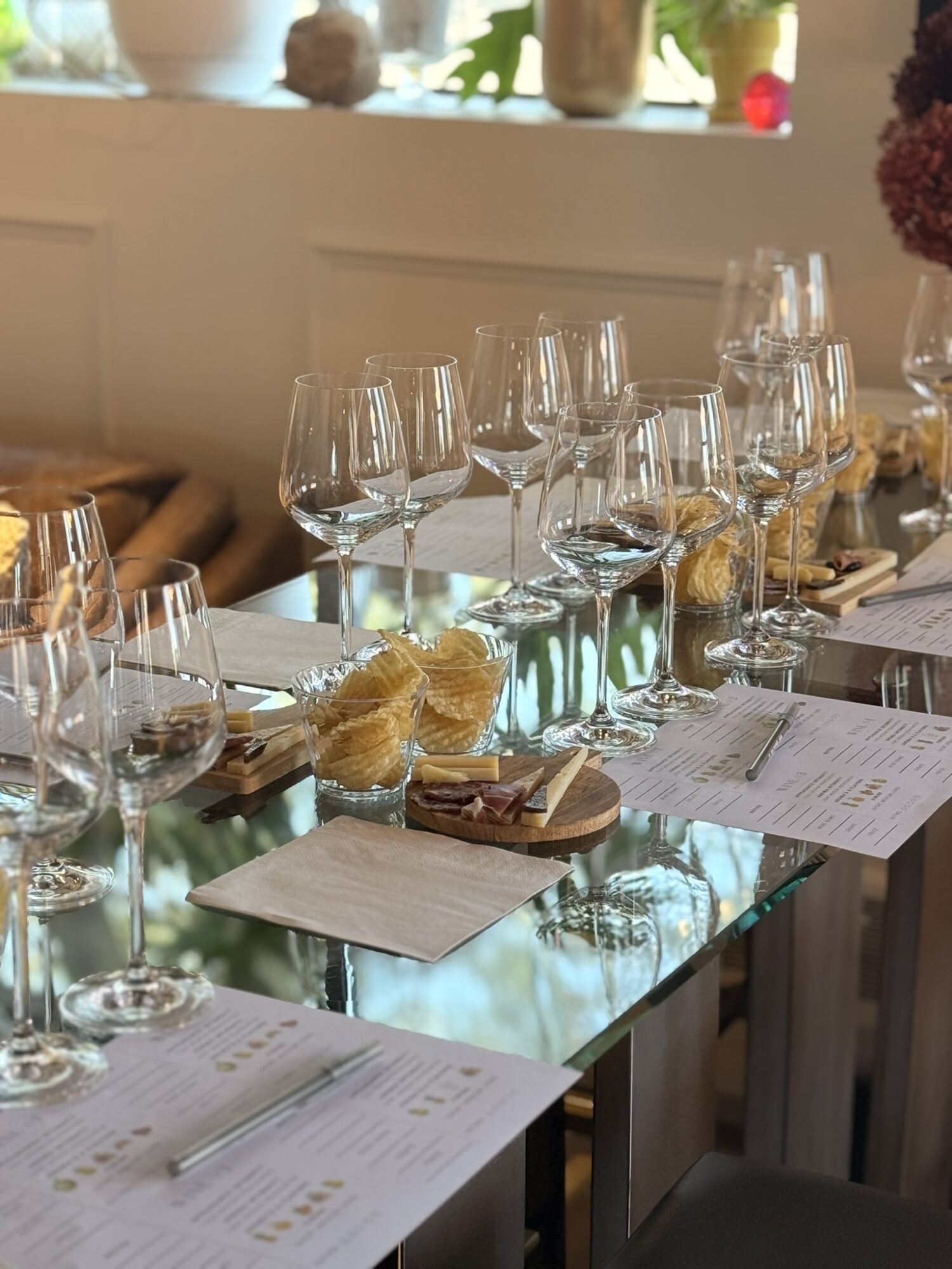 Table set with wine glasses, plates, and napkins, with a window and plants in the background.