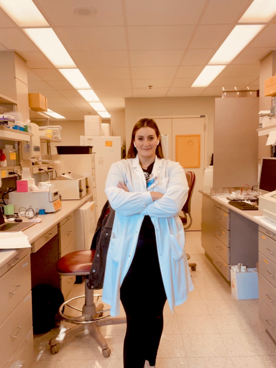 Female scientist in a lab coat standing in a laboratory with equipment and shelves around her.