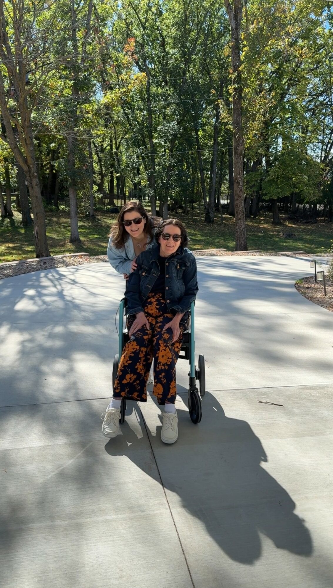 Two women outdoors in a park with trees, one sitting in a wheelchair and the other standing behind her, both wearing sunglasses.