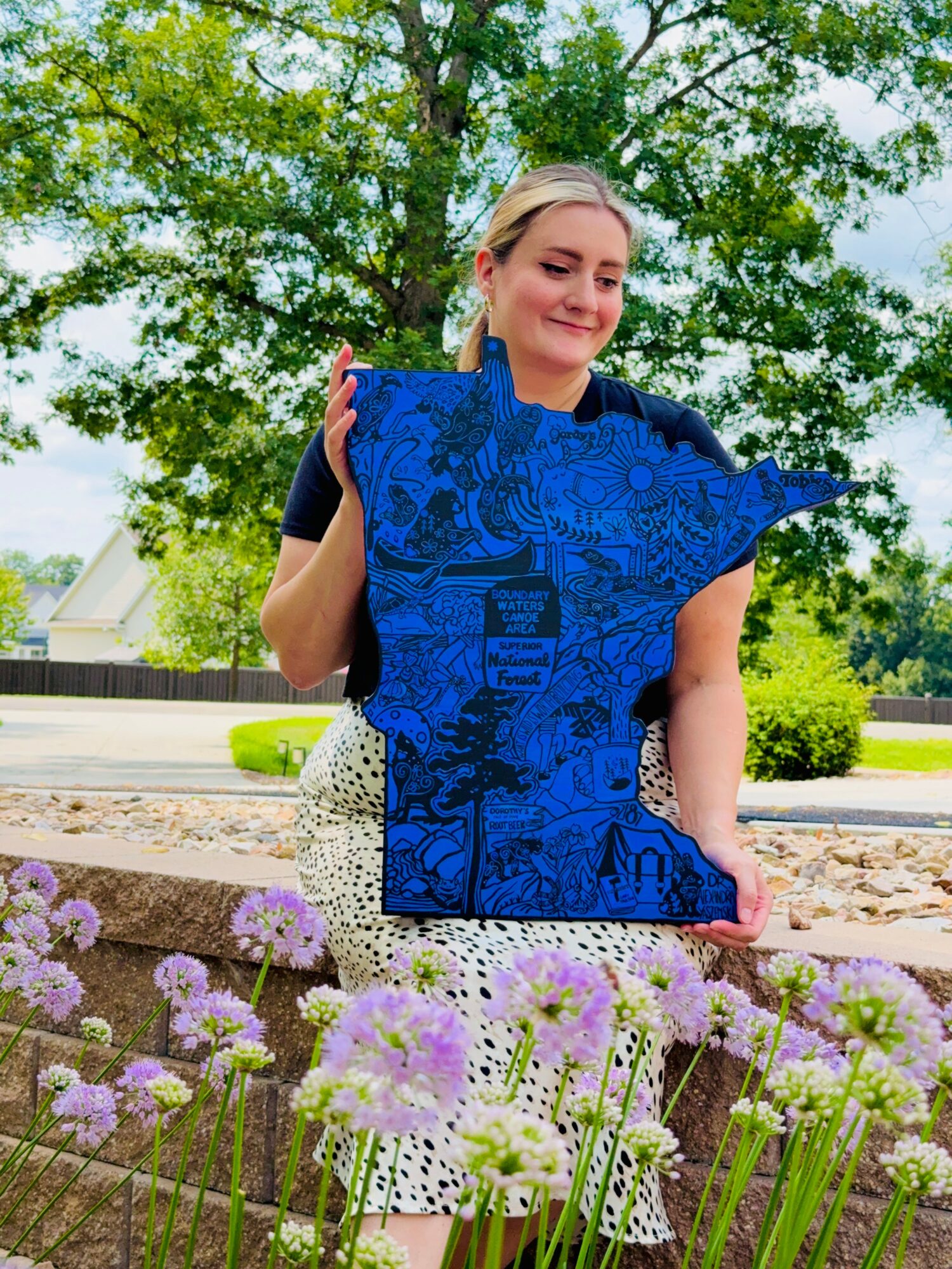 Woman holding a large blue patterned object outdoors with trees and flowers in background.