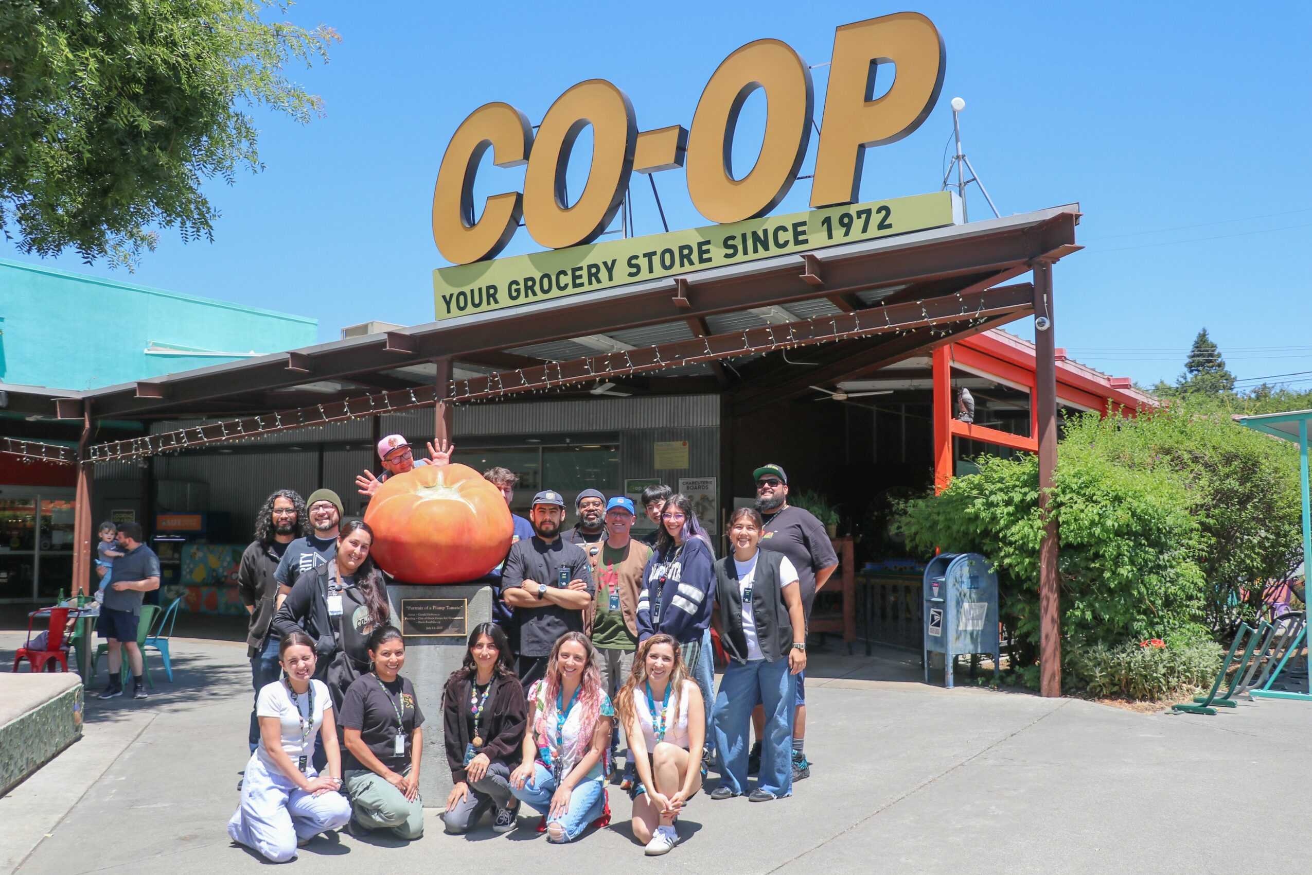 Group of people standing and sitting outside a grocery store with a large pumpkin decoration, trees, and a blue sky.