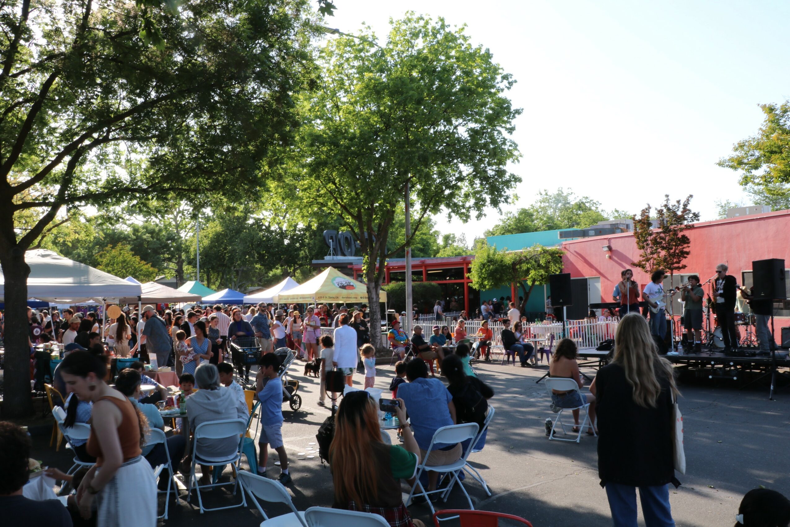 Outdoor event with people, tents, trees, and a stage with performers, under a clear sky.
