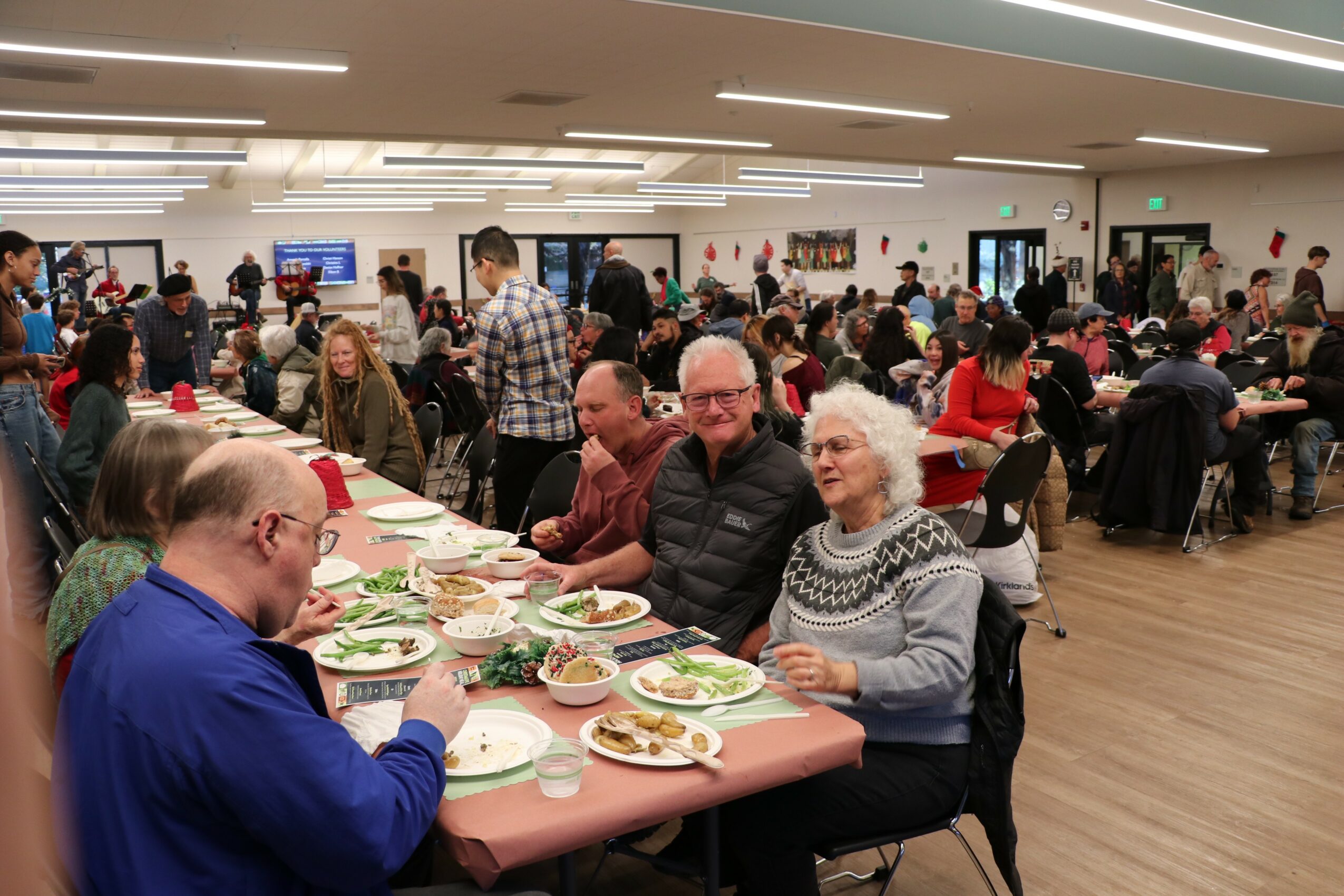 People dining at tables in a large, well-lit room with many guests and servers, some standing and some seated, enjoying a meal.