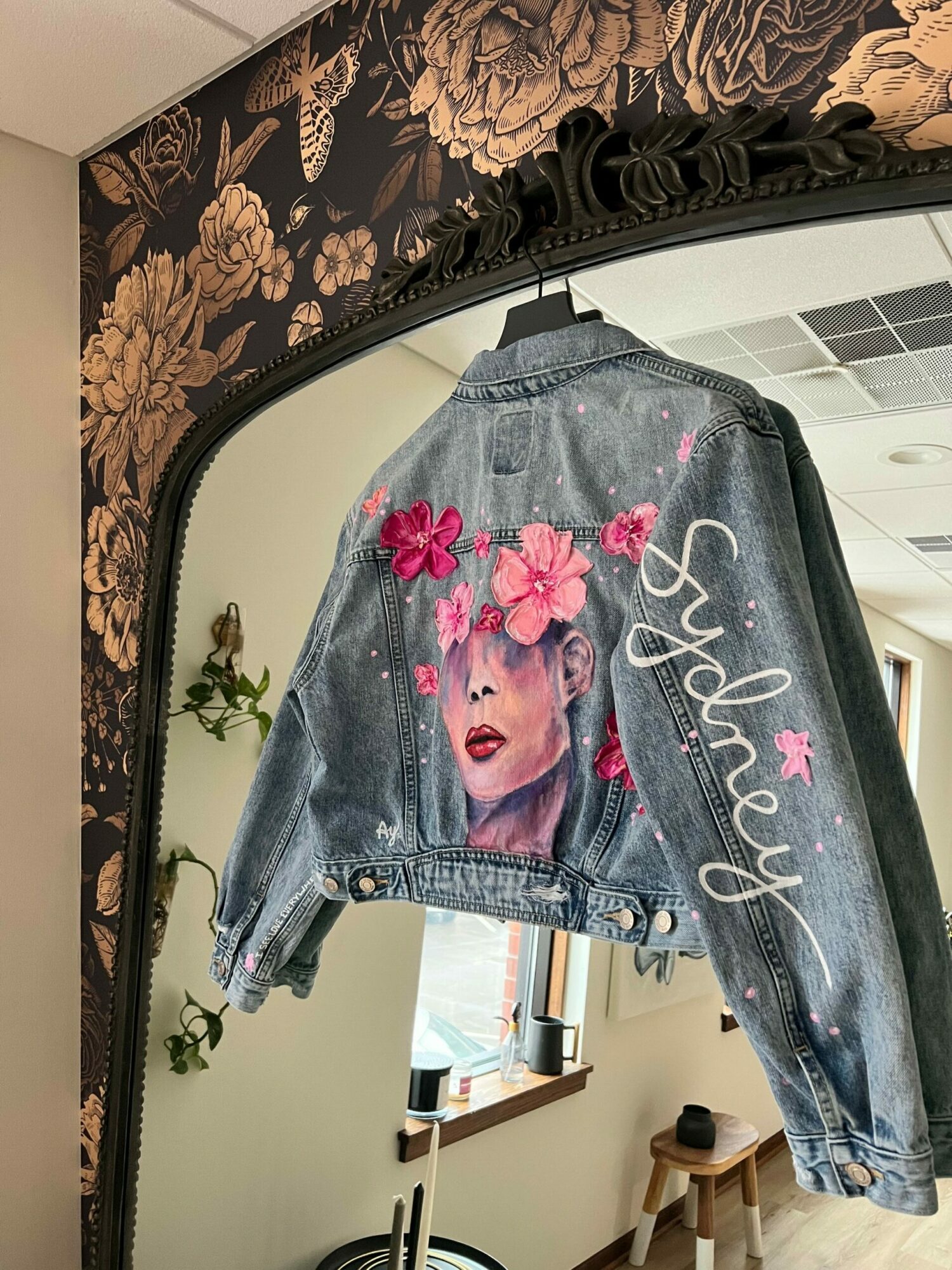 Denim jacket with pink floral design and the name 'Sydney' written on it, hanging on a mirror. Word count: 20.