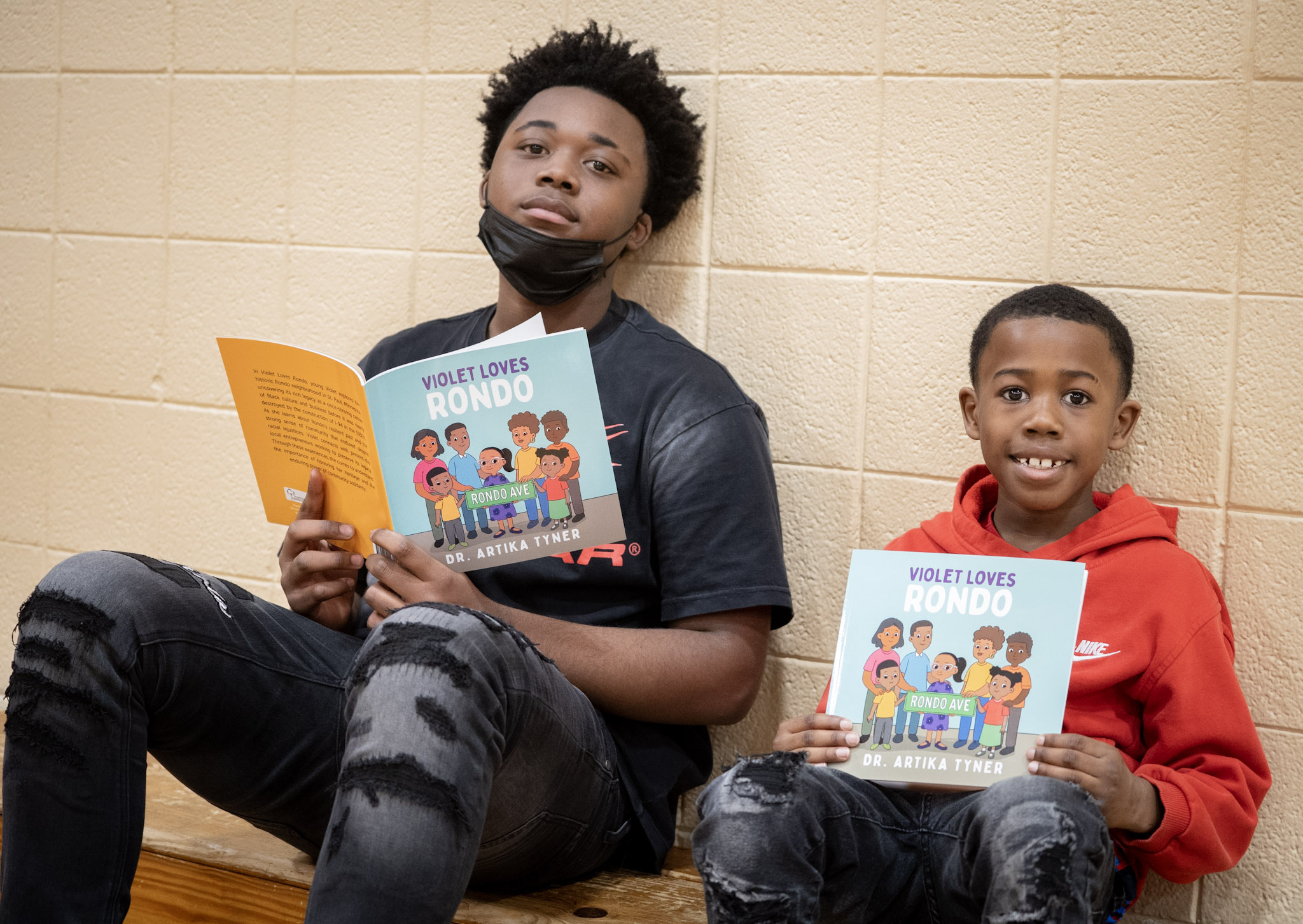 Two children sitting against a beige wall, holding books titled 'Colectivos Rondo,' smiling and reading.