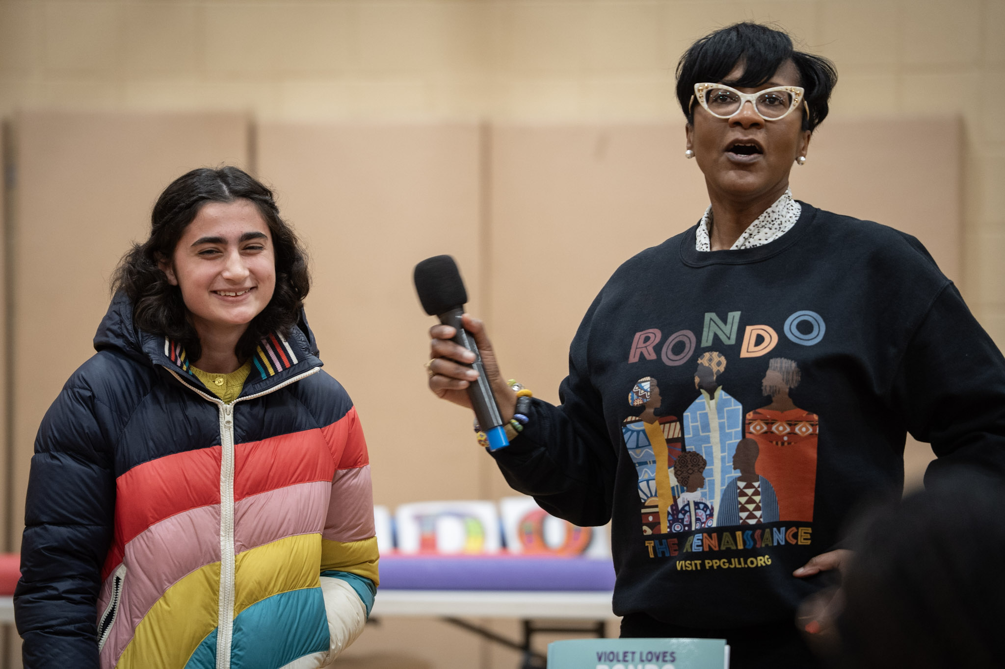 Two women speaking at an event, one holding a microphone, with a table and beige wall in background.