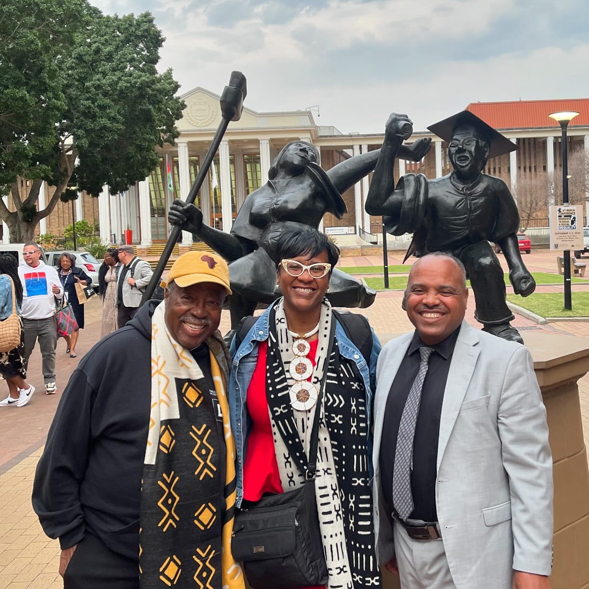 Three people smiling in front of a statue with two figures holding a paddle and a graduation cap, in an outdoor setting.