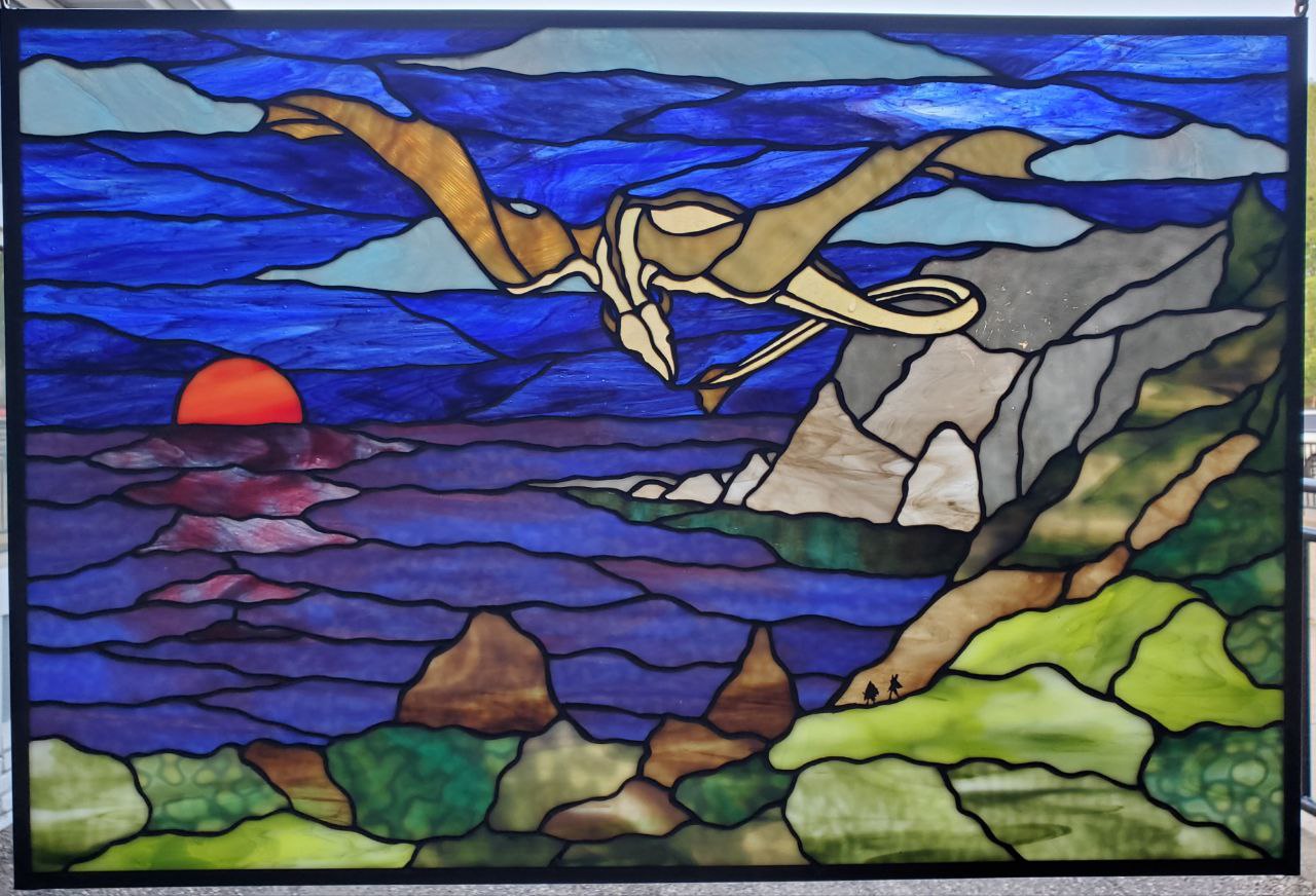 Stained glass window depicting a bird flying over a landscape with a setting sun, rocks, and greenery.