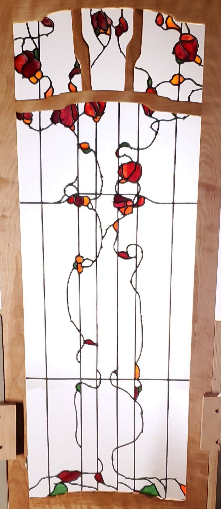 Vertical stained glass window with red and orange flowers and green leaves, featuring black wire-like outlines.