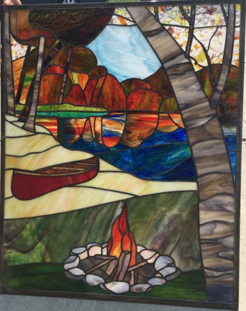 Colorful stained glass window depicting a landscape with trees, a river, and a campfire with rocks.
