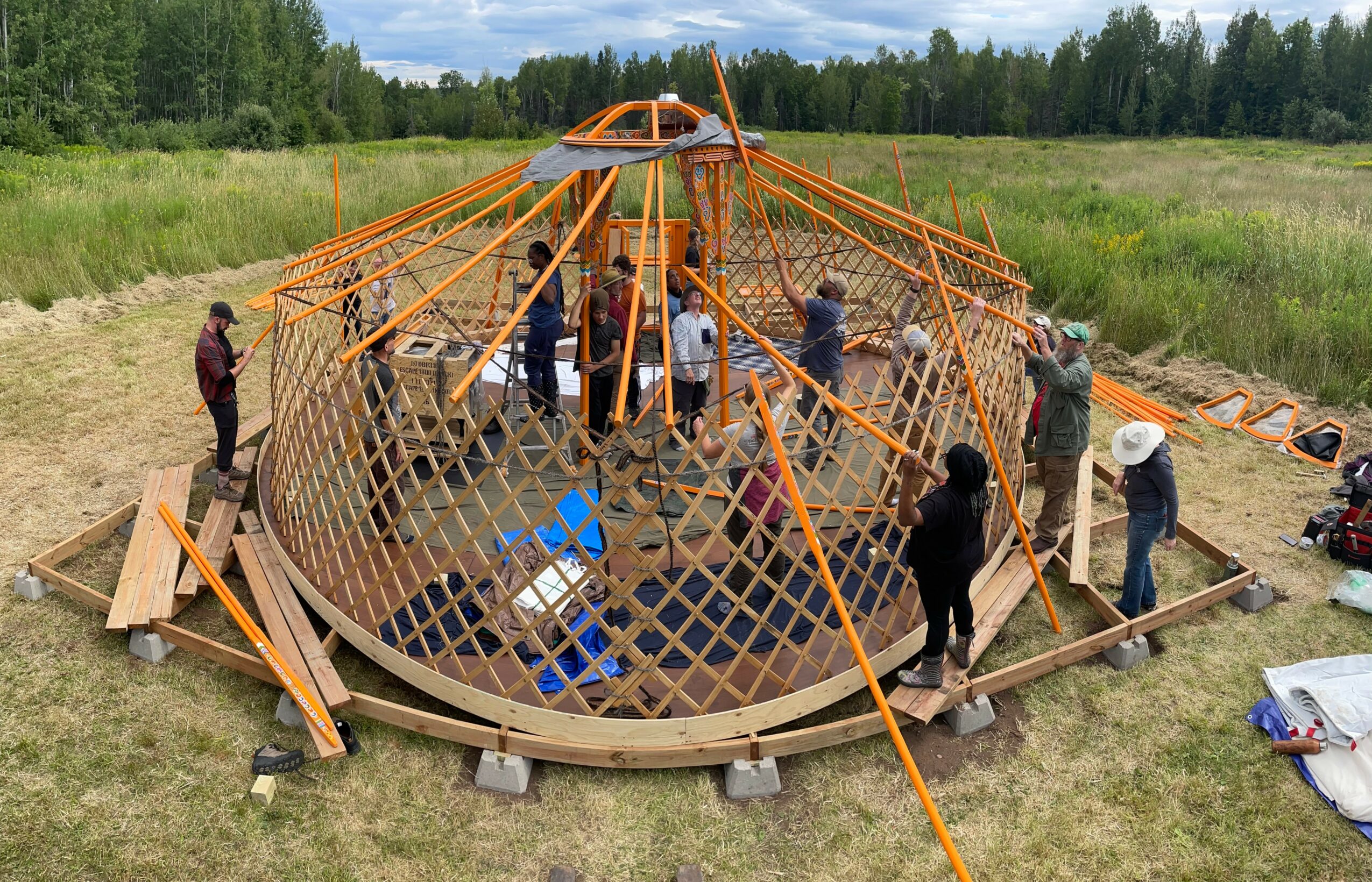 Group of people assembling a large, circular, orange rope structure outdoors in a grassy field with trees in the background.