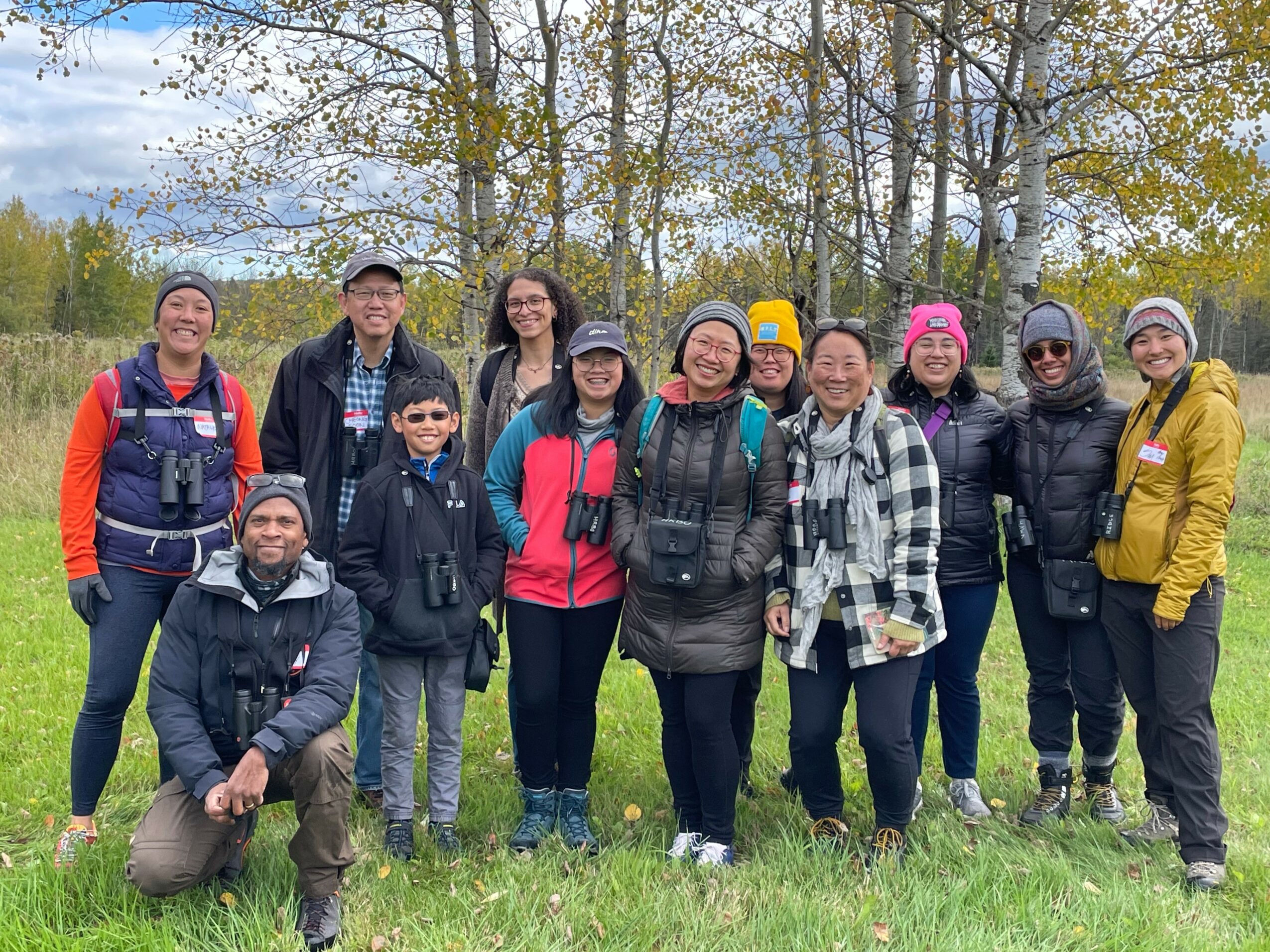 Group of ten people outdoors in a grassy area with trees and cloudy sky, smiling and posing for a photo.