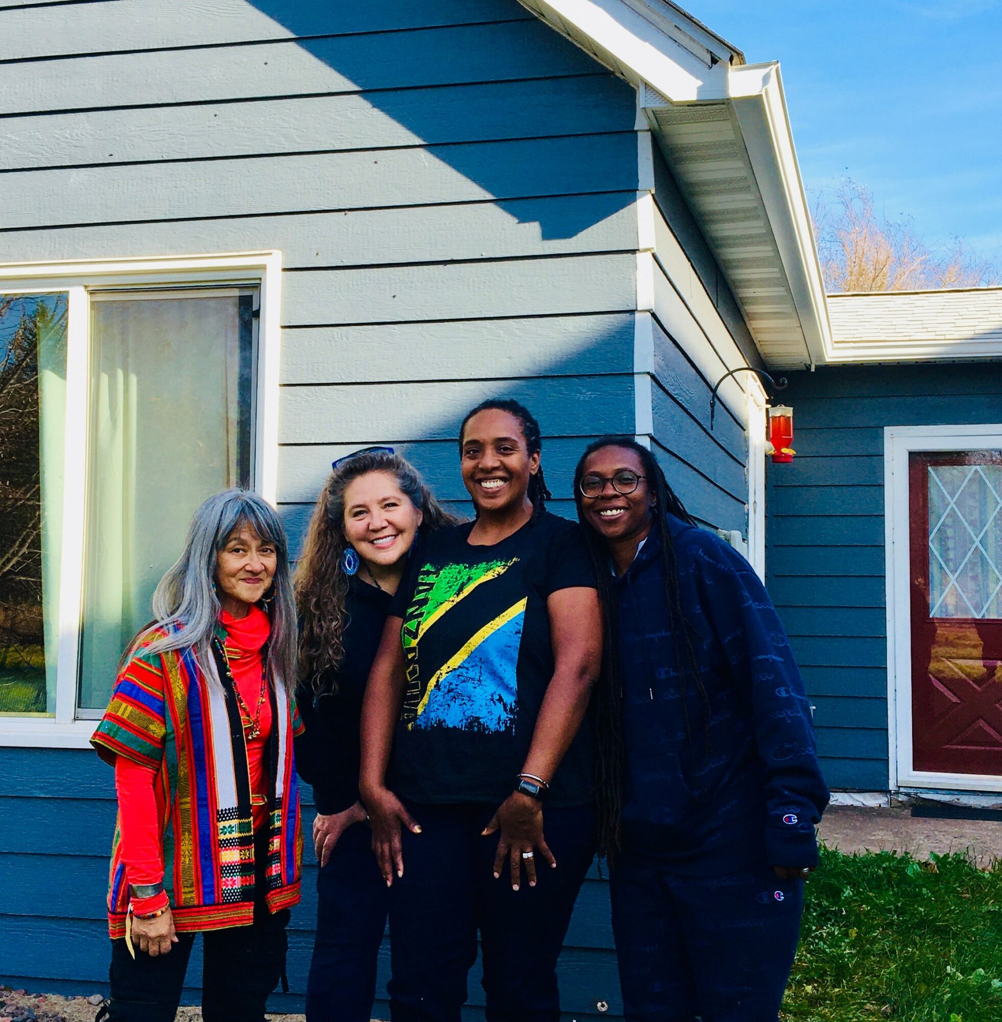 Four women standing outside a blue house, smiling, with a window and door visible, bright sky above.