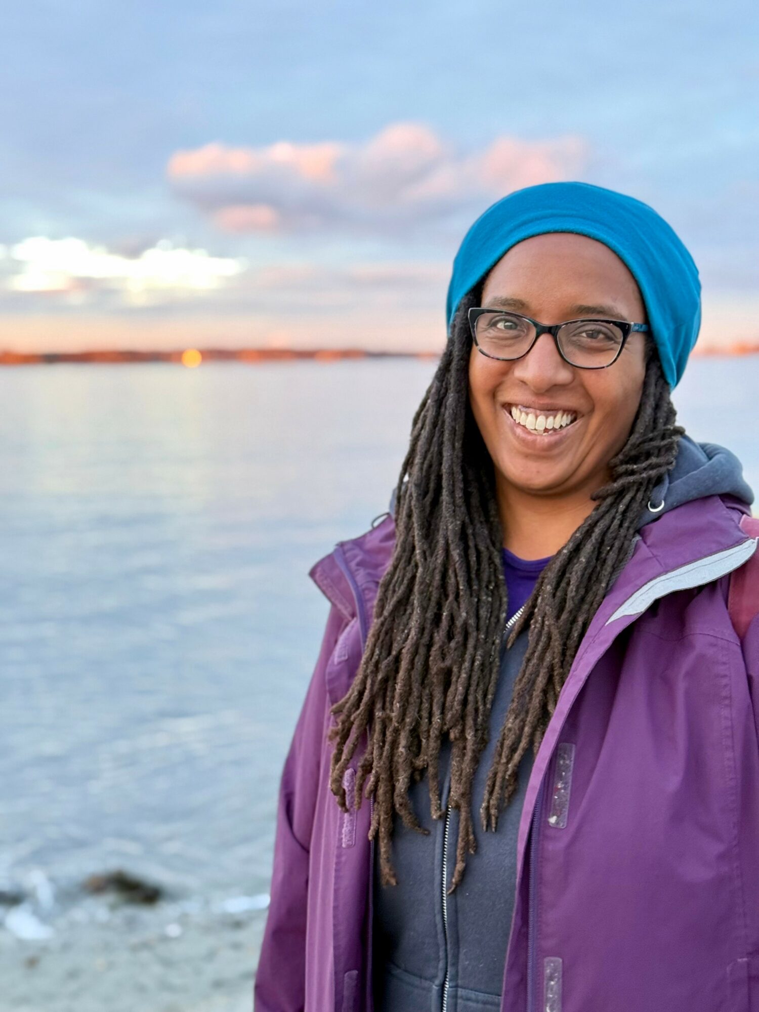 Smiling woman with glasses and dreadlocks wearing a blue headscarf and purple jacket outdoors near water.