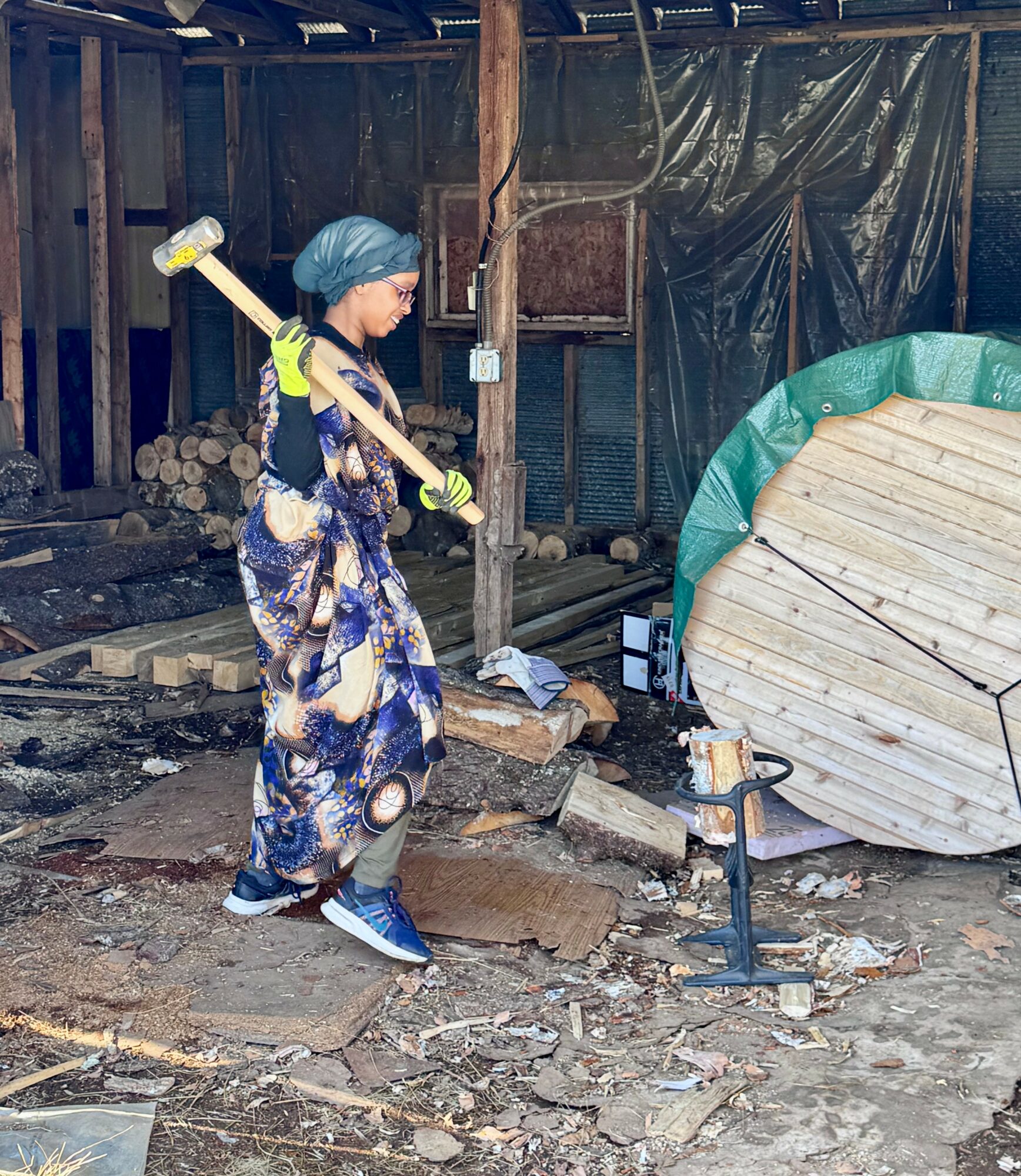 Person wearing a blue headscarf and patterned pants swings a hammer outdoors on uneven ground with wood and construction materials nearby.