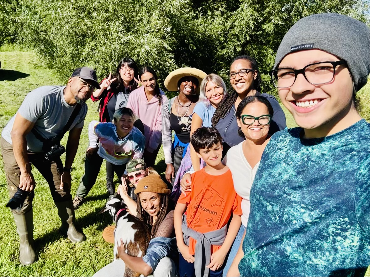 Group of people outdoors on grass, smiling, posing for a photo, with trees in background.