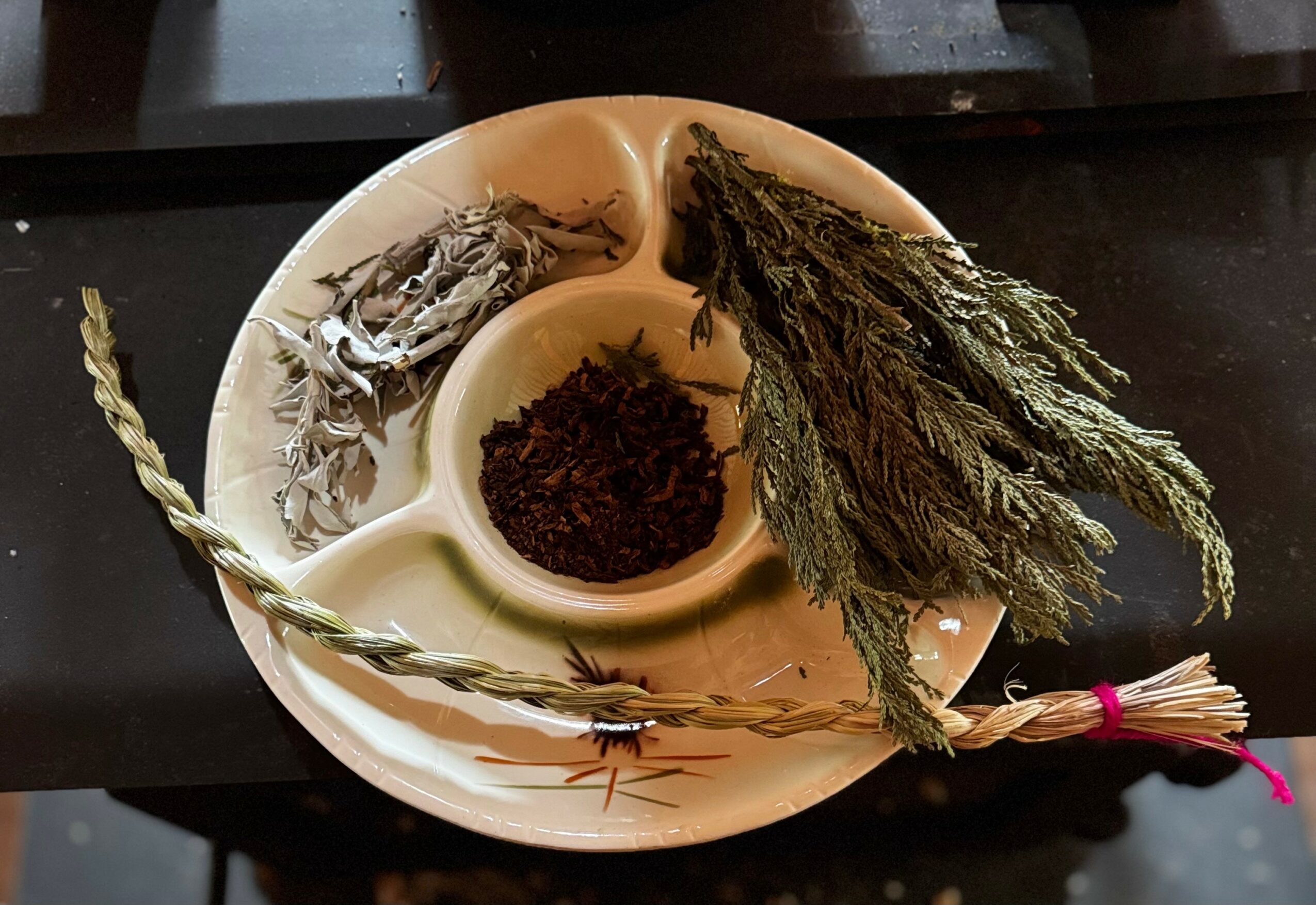 A divided dish with dried herbs, seaweed, and a twisted stick, on a dark surface.