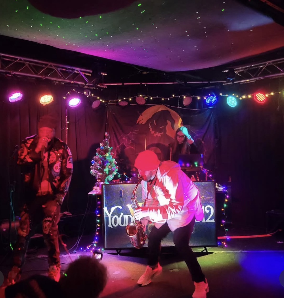 Two performers on stage with colorful lights, a Christmas tree, and a DJ setup, one playing saxophone and the other dancing.