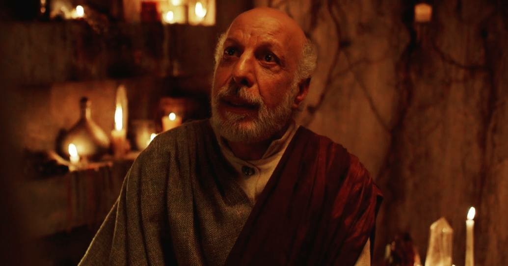 A man with a beard and bald head looks upward in a dimly lit room with candles and stone walls.