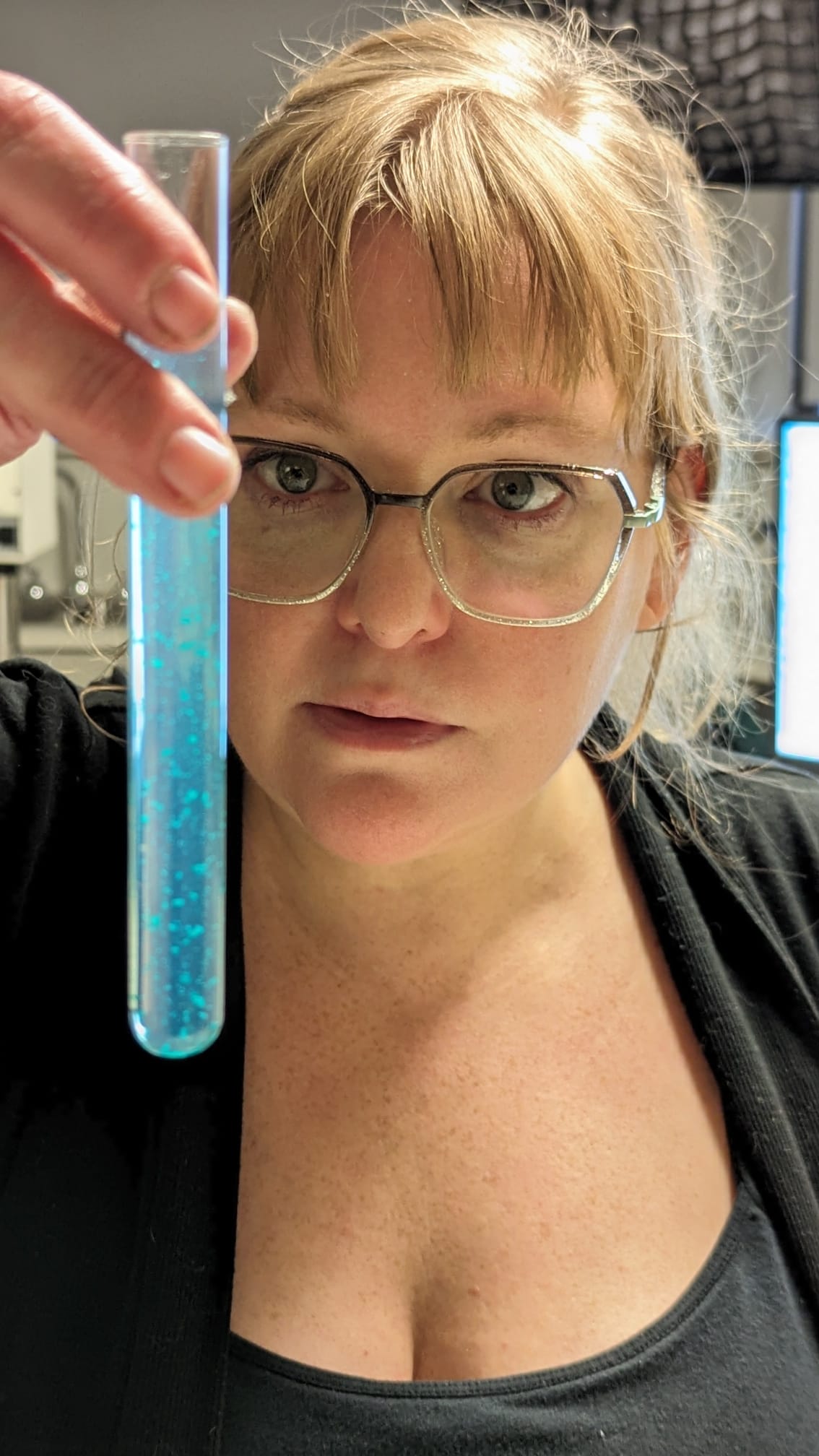 Woman with glasses holds a test tube with blue liquid, looking at it intently in a laboratory setting.