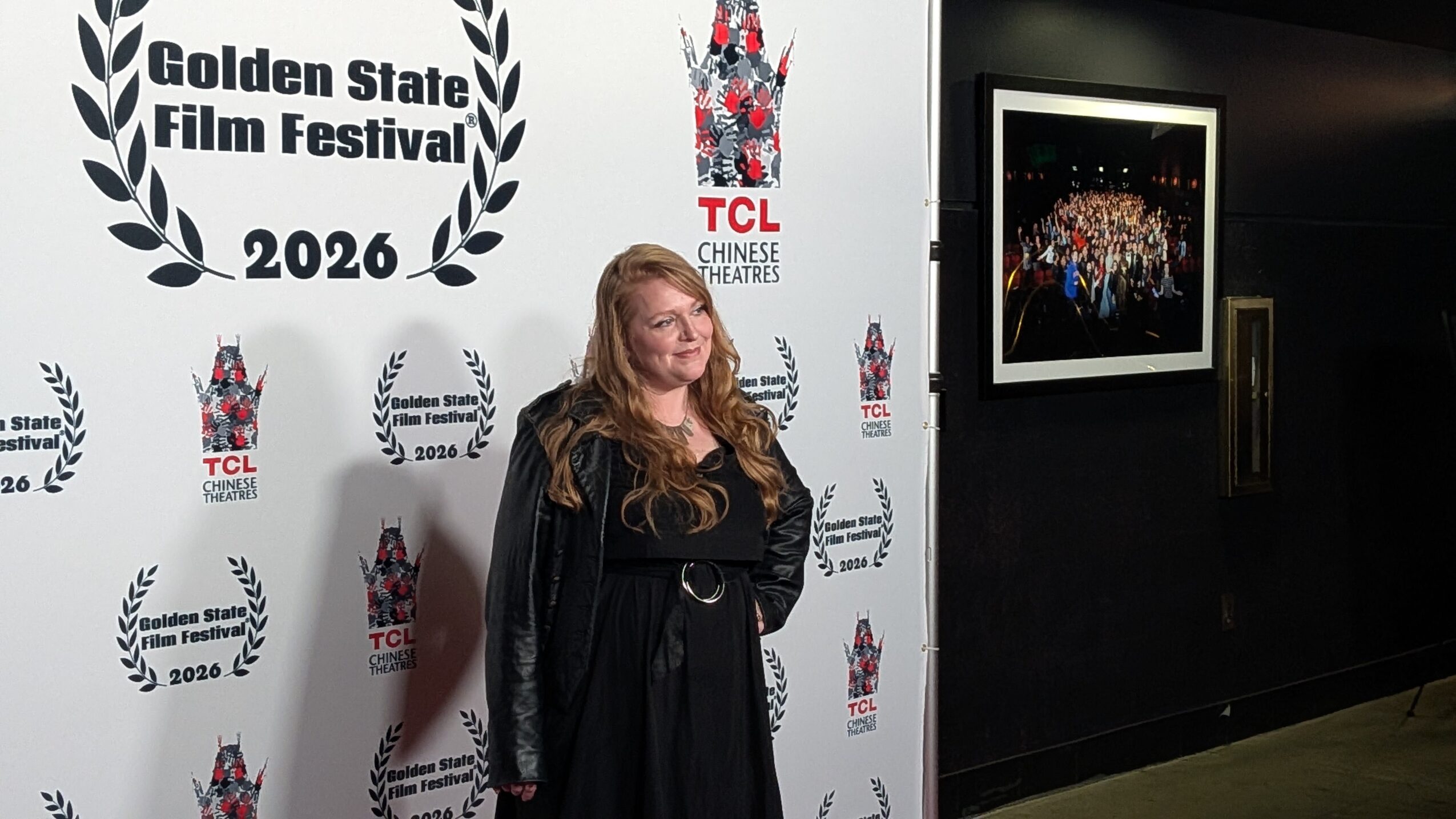 Woman with long hair in black dress standing in front of a white backdrop with festival logos and text.