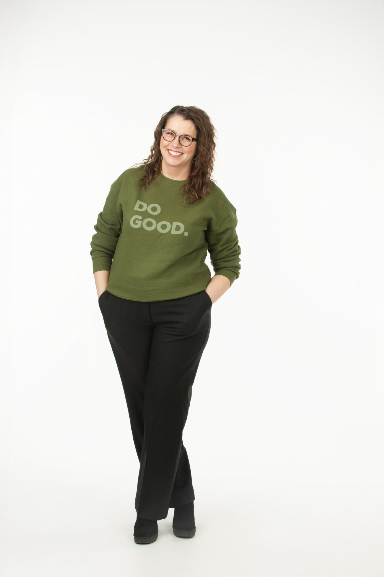 Woman with curly hair and glasses smiling, wearing a green sweater with 'DO GOOD.' text, black pants, standing with hands in pockets.