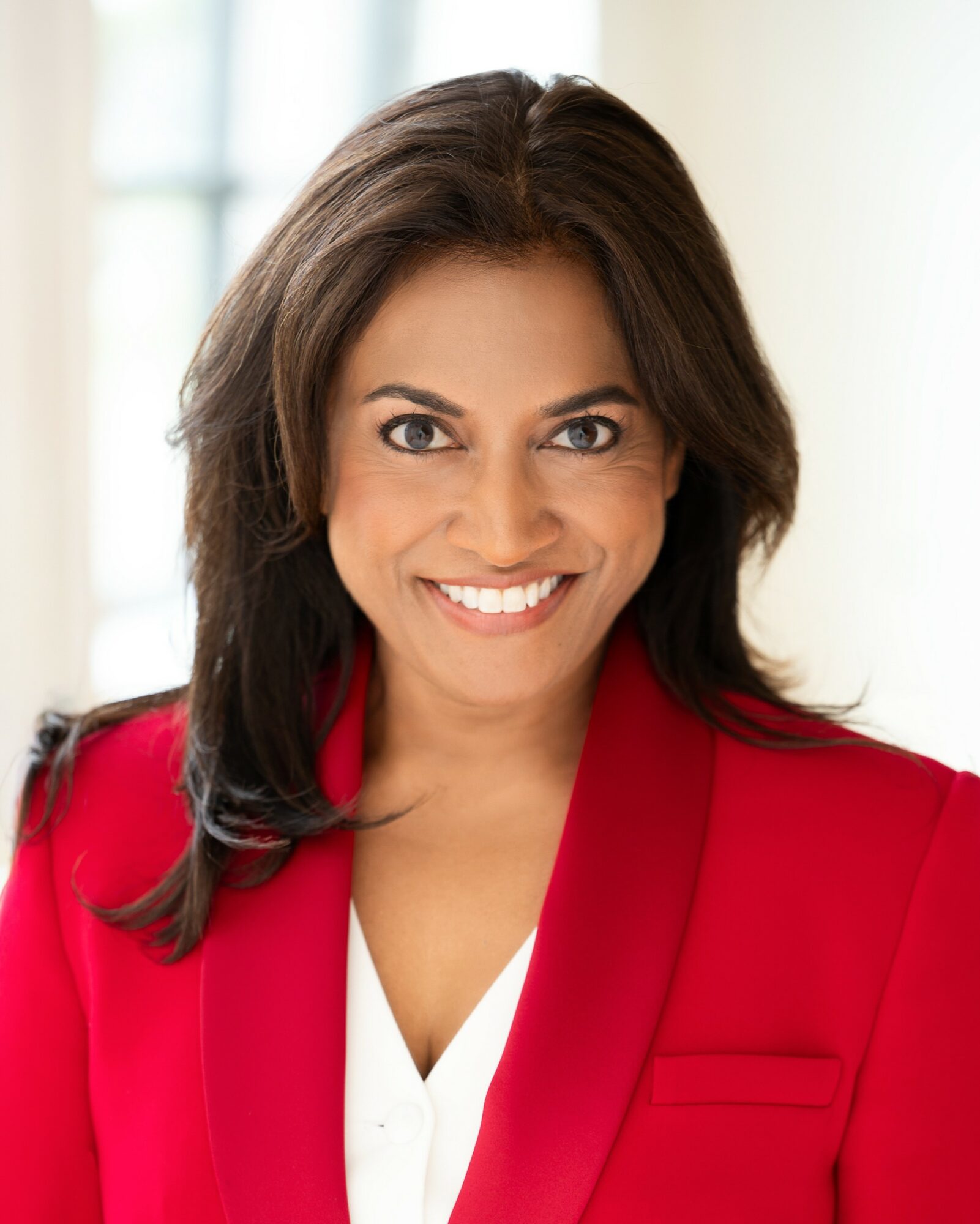 Woman with shoulder-length dark hair smiling, wearing a red blazer and white top, in a bright indoor setting.